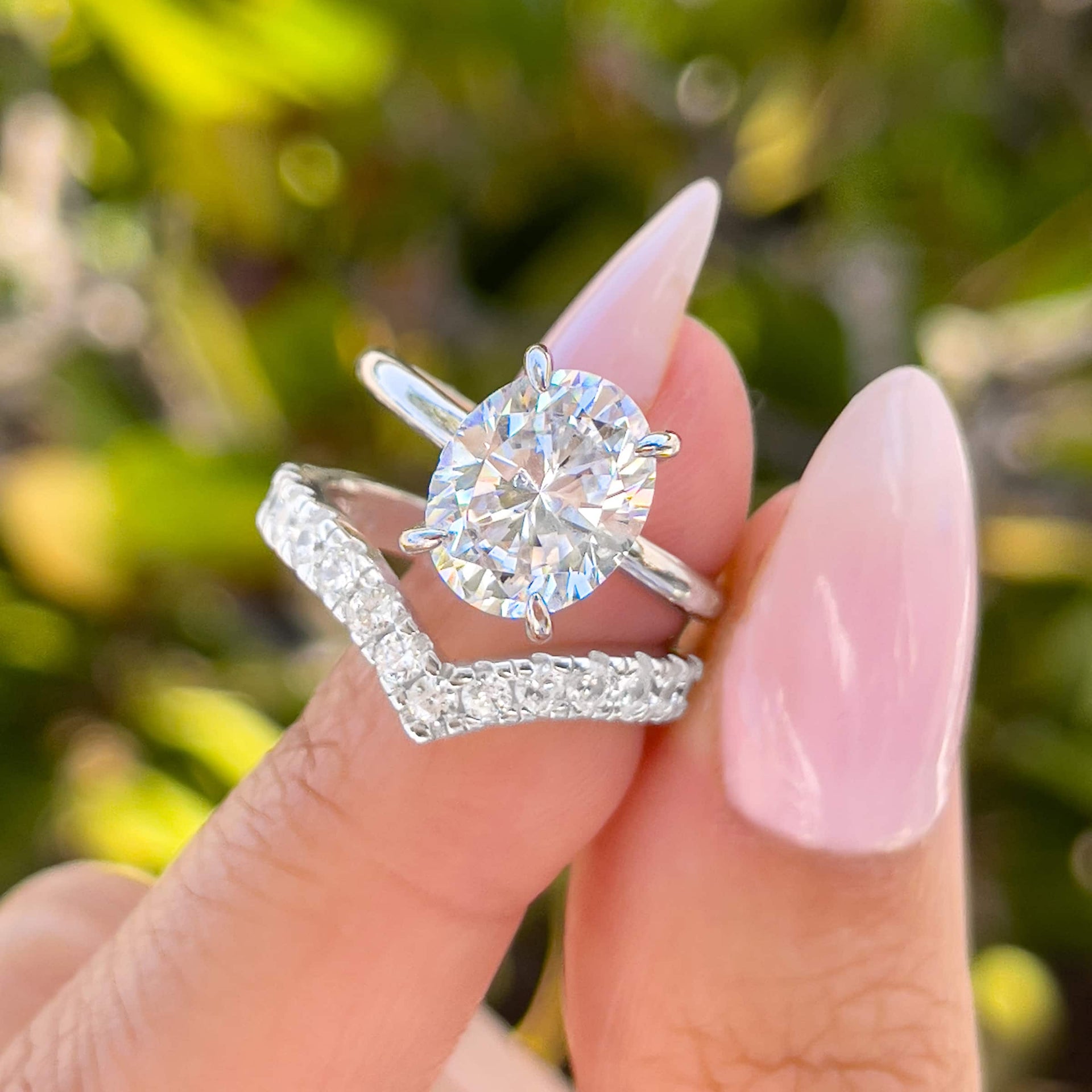 stunning silver oval cut wedding set and thick chevron wedding band held by model with light pink nails in front of greenery