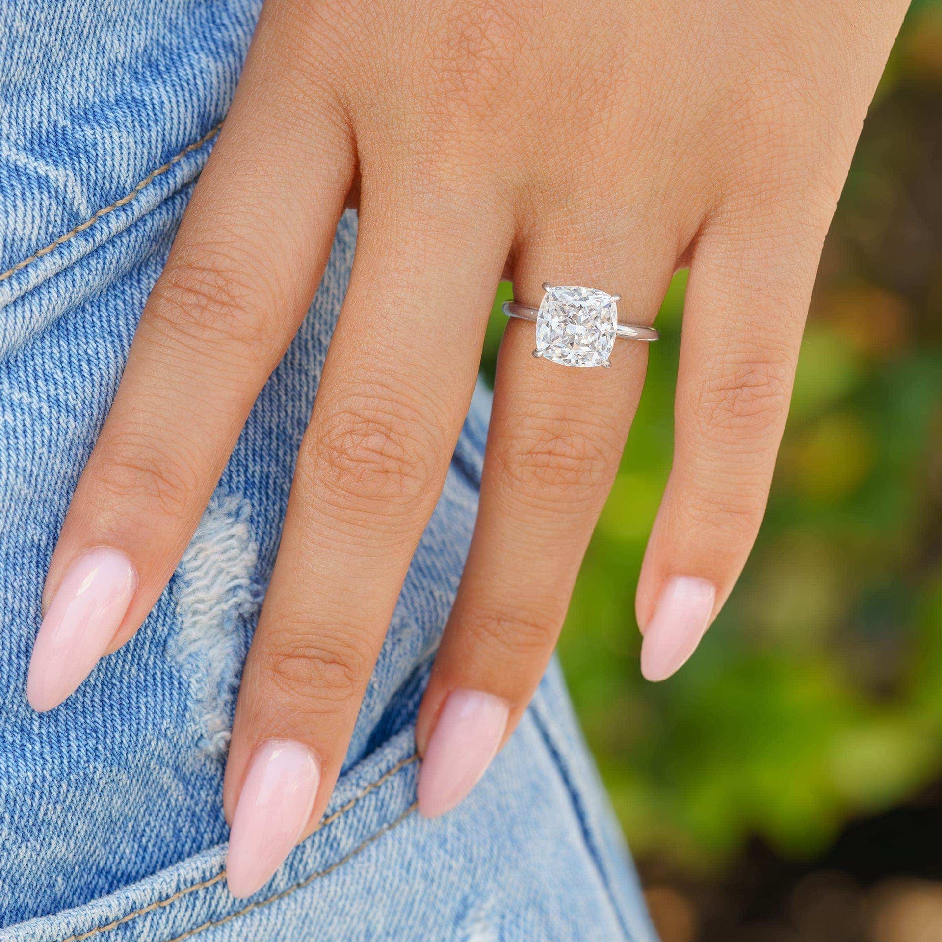 The Claire silver engagement ring with cushion-cut simulated diamond solitaire shown on hand in natural lighting with pink nails and denim background