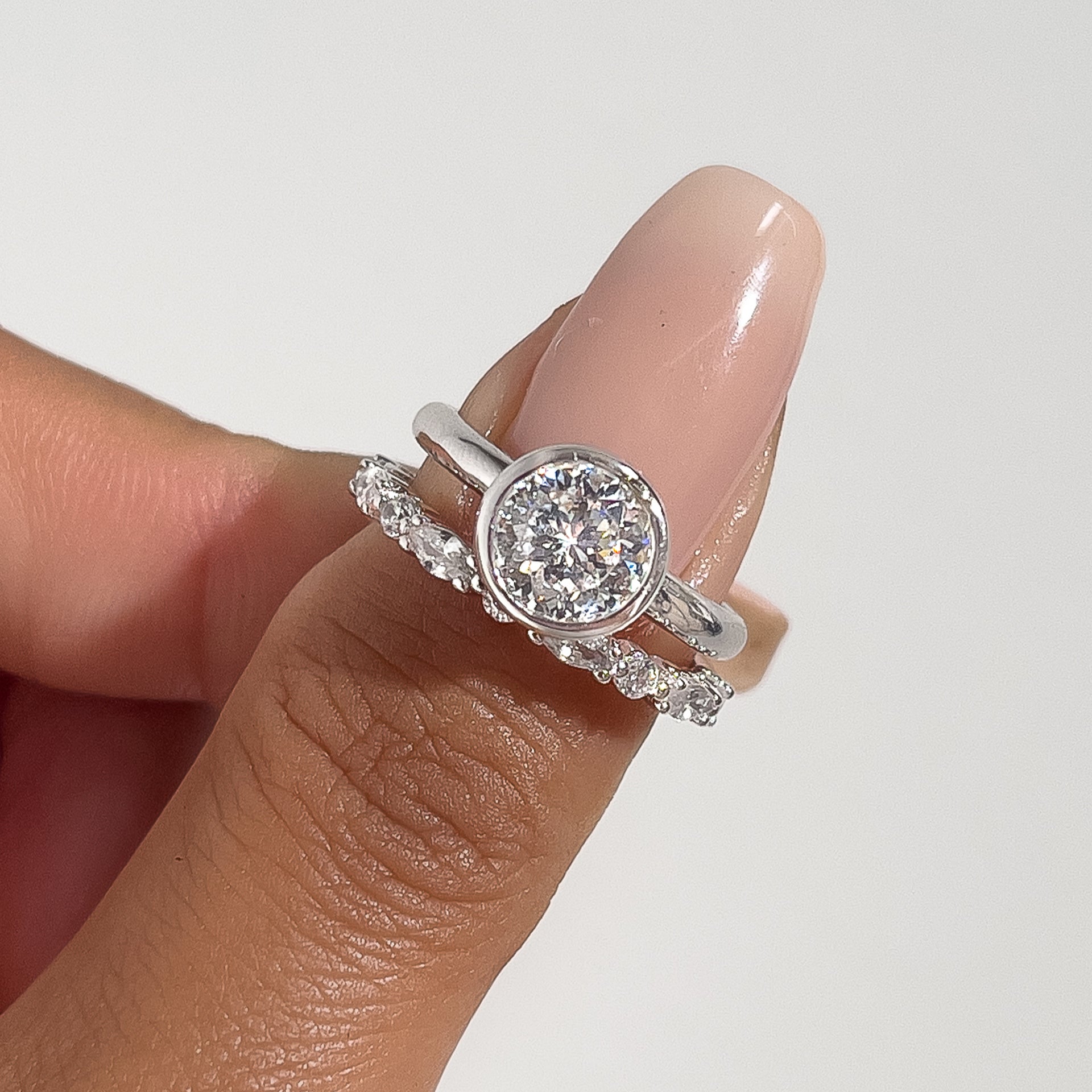 a close-up shot of a silver bezel round cut ring with a pave silver wedding band on model's thumb with pink manicured nails