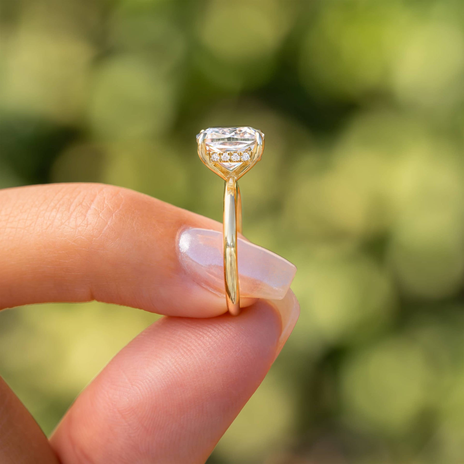 Close-up of The Claire gold engagement ring with cushion-cut simulated diamond solitaire and hidden halo detailing, held between fingers in natural light