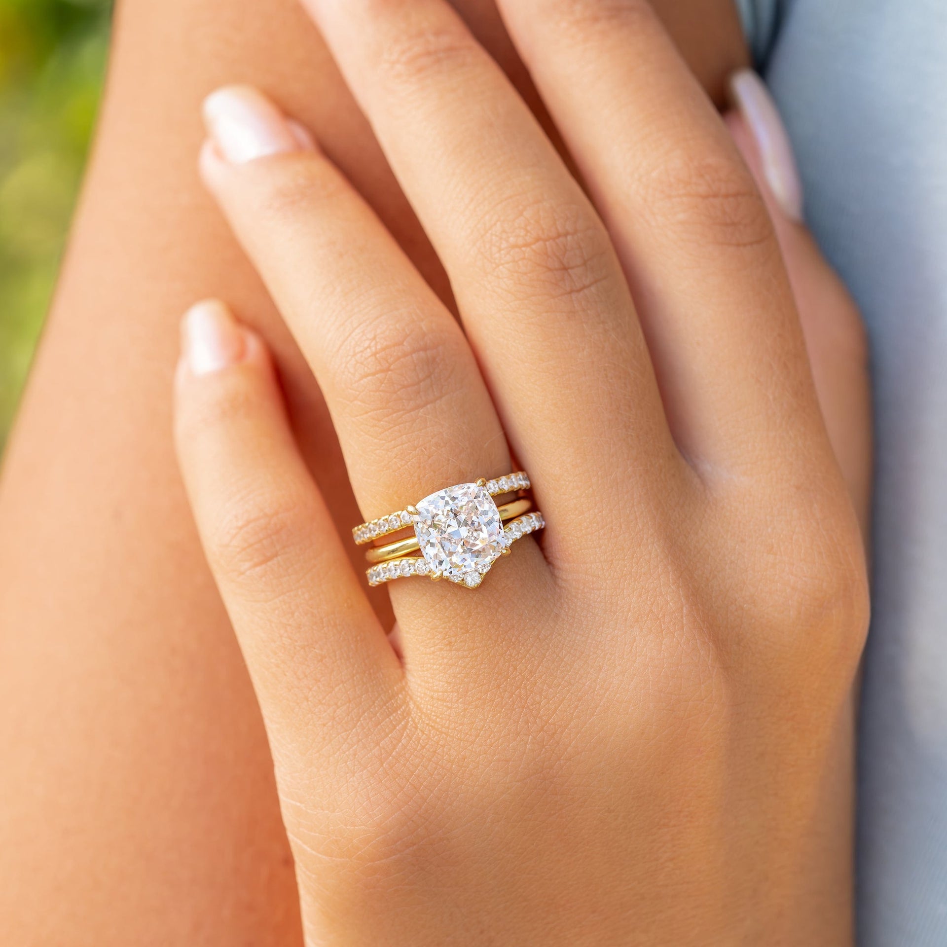The Claire cushion-cut solitaire engagement ring in a yellow gold setting stacked between two pavé bands, shown on hand in natural light