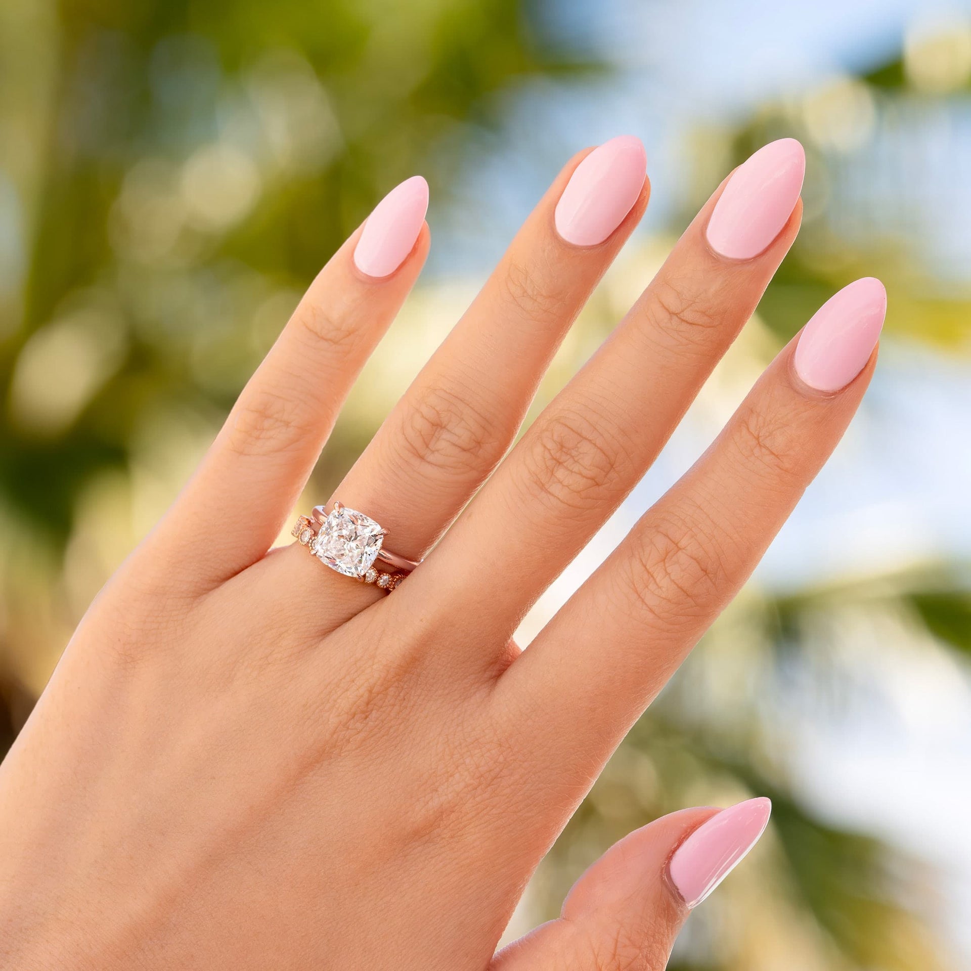 The Claire rose gold engagement ring with cushion-cut simulated diamond solitaire shown on hand with pink almond nails in bright natural light