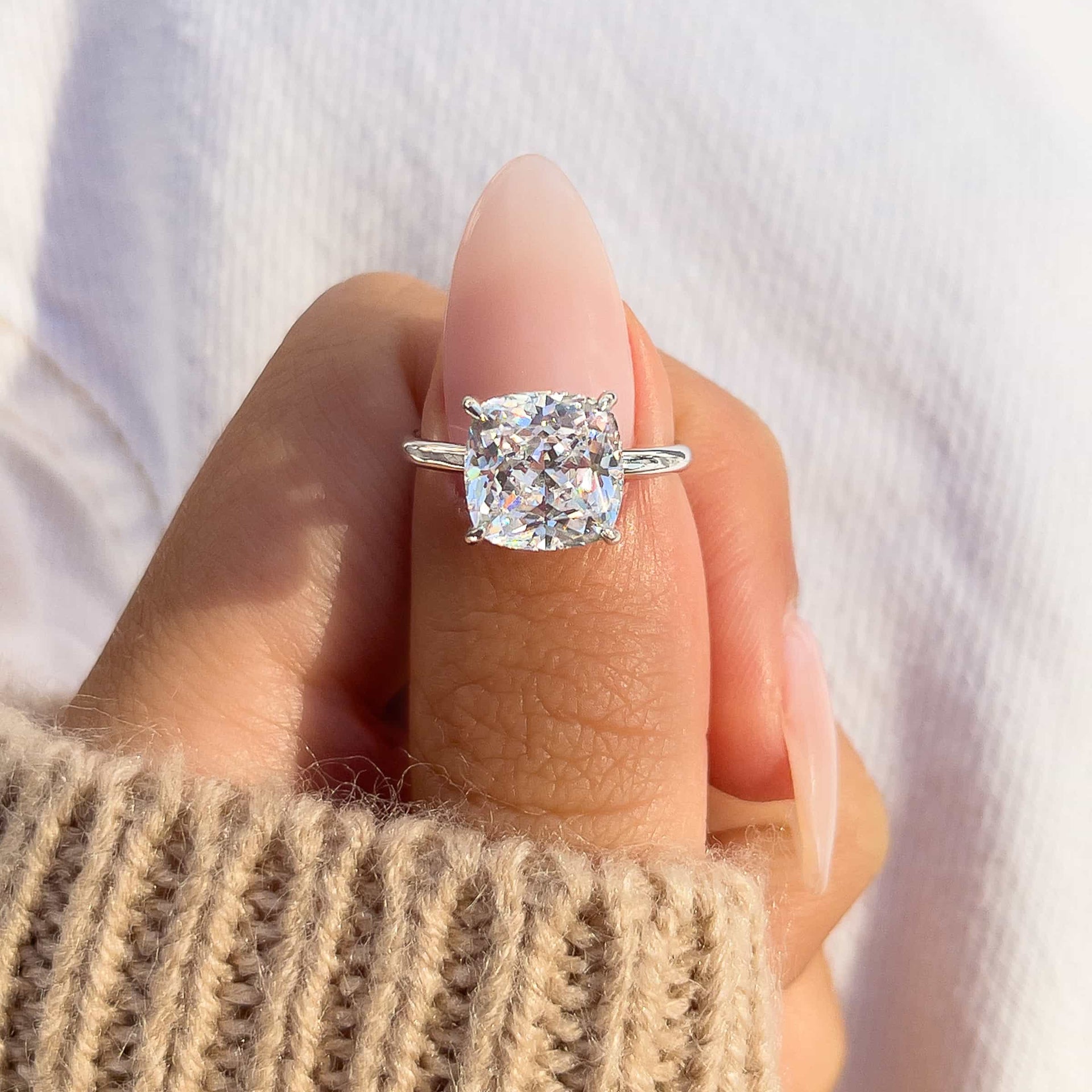Close-up of The Claire silver engagement ring with 3.5 carat cushion-cut simulated diamond solitaire, held on thumb with neutral manicure