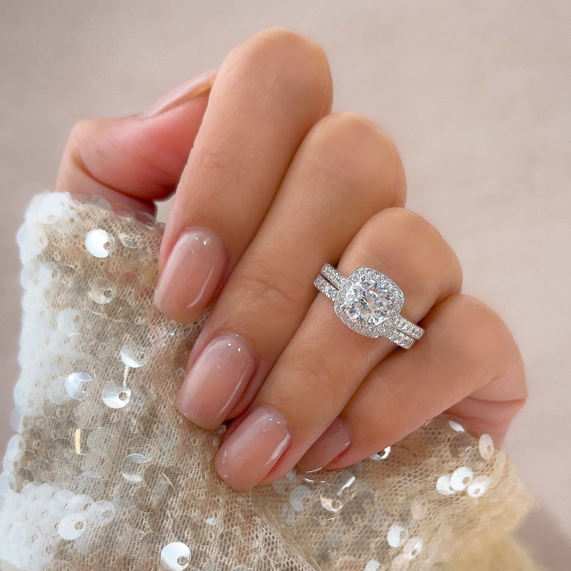 Close-up of The Evermore silver ring set with 1.5 carat round-cut simulated diamond in cushion halo and pavé band, with neutral nails and sequin sleeve