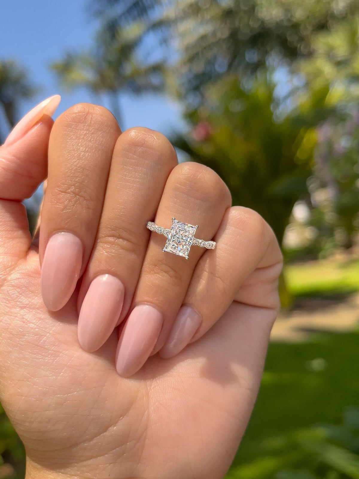 A hand with pink nails is held up against a green outdoor background, wearing a silver engagement ring with a rectangular center stone and a stone-lined band.