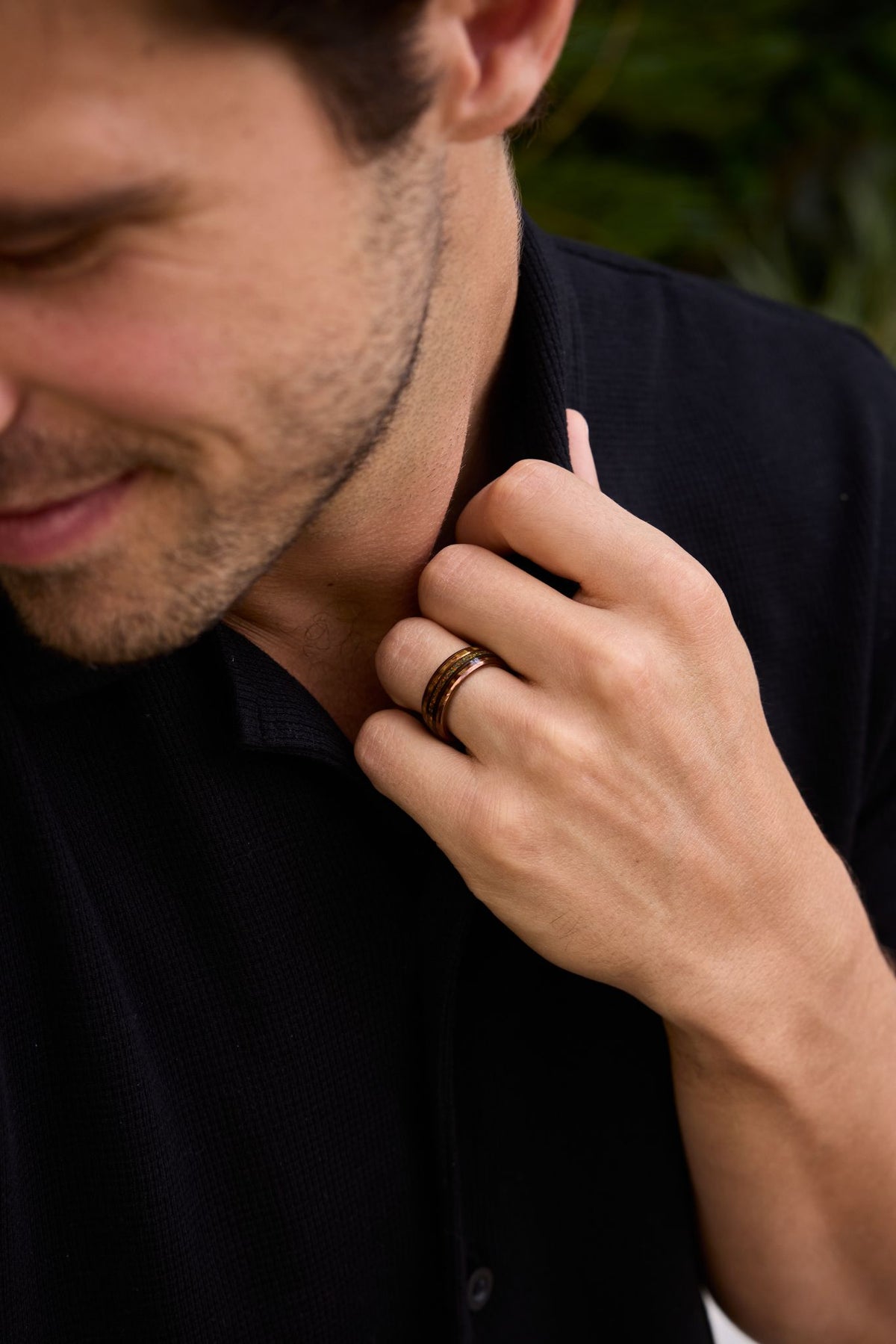 A person in a dark shirt looks down with a slight smile while touching their collar, wearing a thin band on one finger.