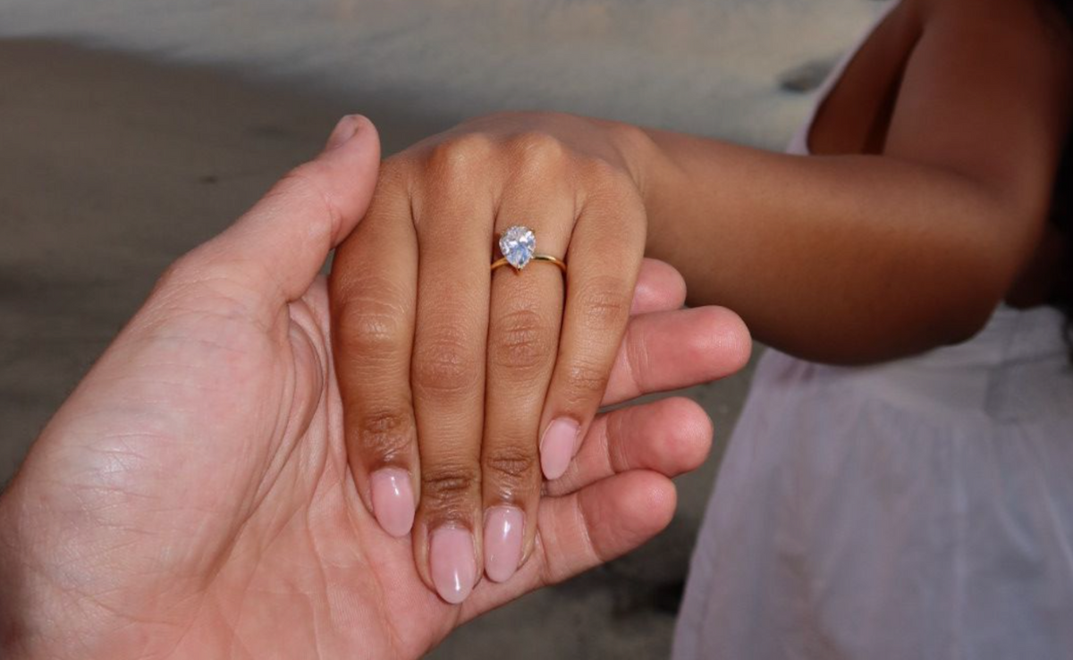 Two people hold hands on a beach at sunset, with one of them wearing a simulated diamond engagement ring.