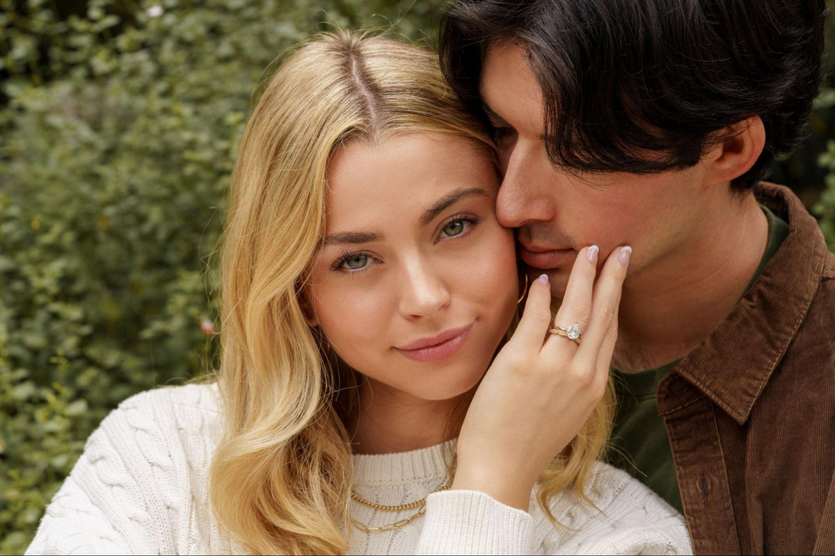 A couple posing cheek to cheek in front of green foliage, with a woman in a cream cable-knit sweater resting a hand on her partner’s face to show a pear-shaped engagement ring on a gold band.