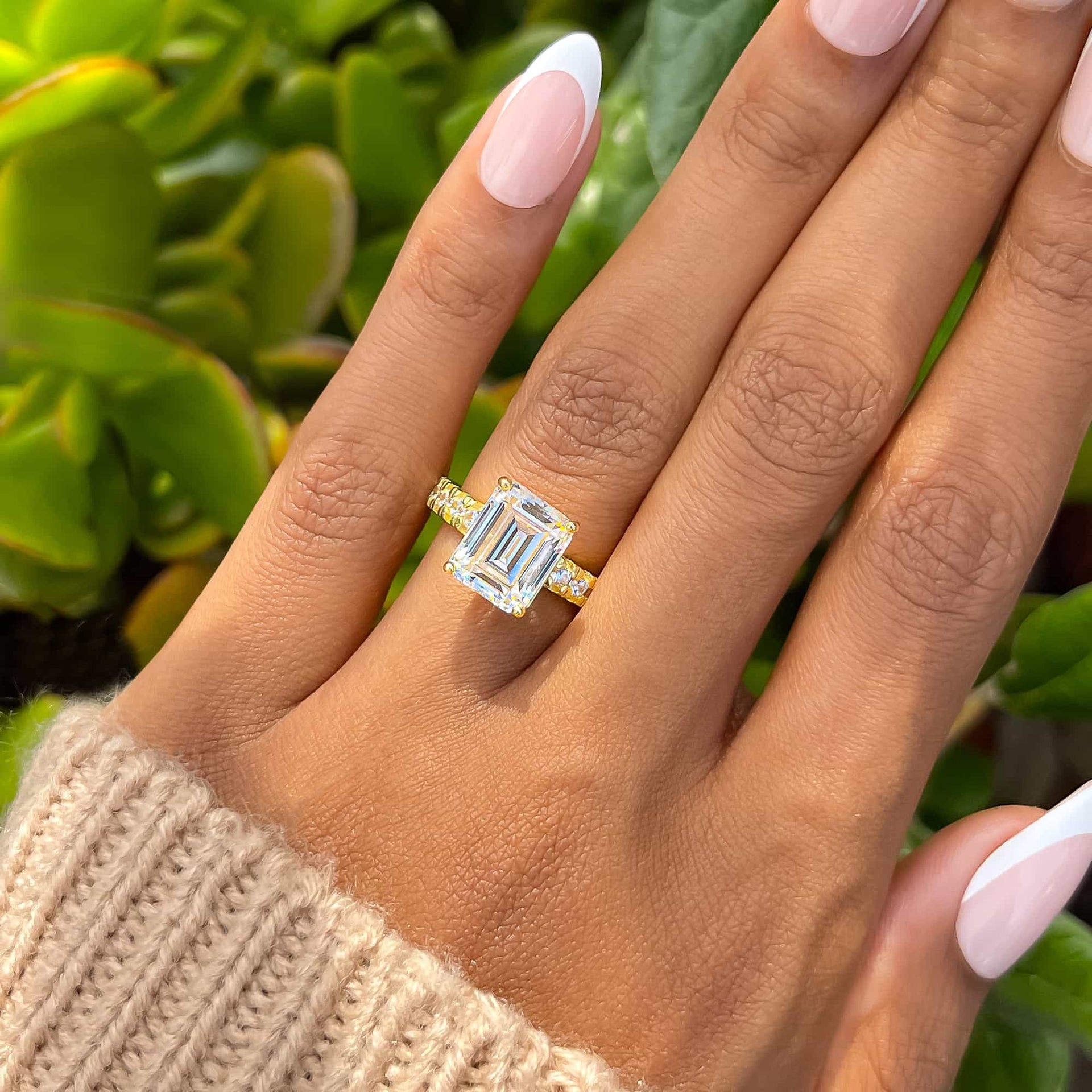 model with french tip nails wearing a gold 3.75 carat emerald cut engagement ring and a tan sweater