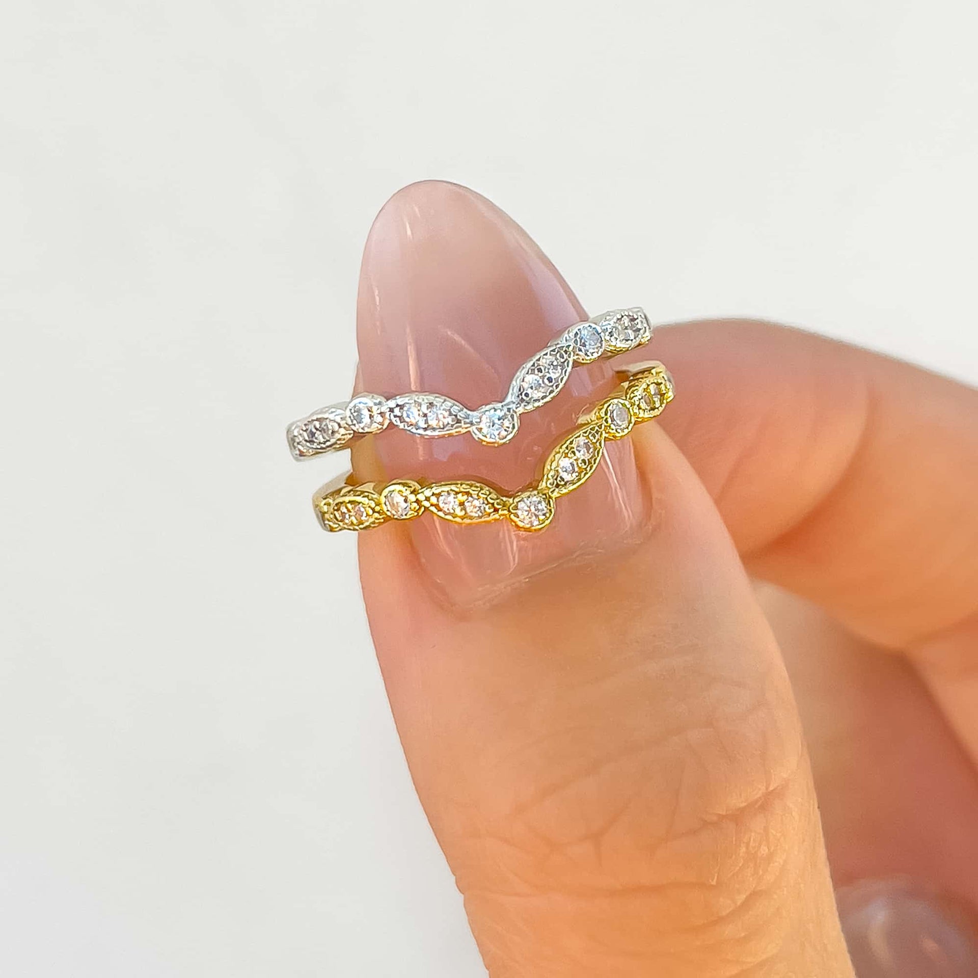 a gold and a silver vintage style chevron wedding band on a model's thumb with neutral pink nails and a white wall in the background