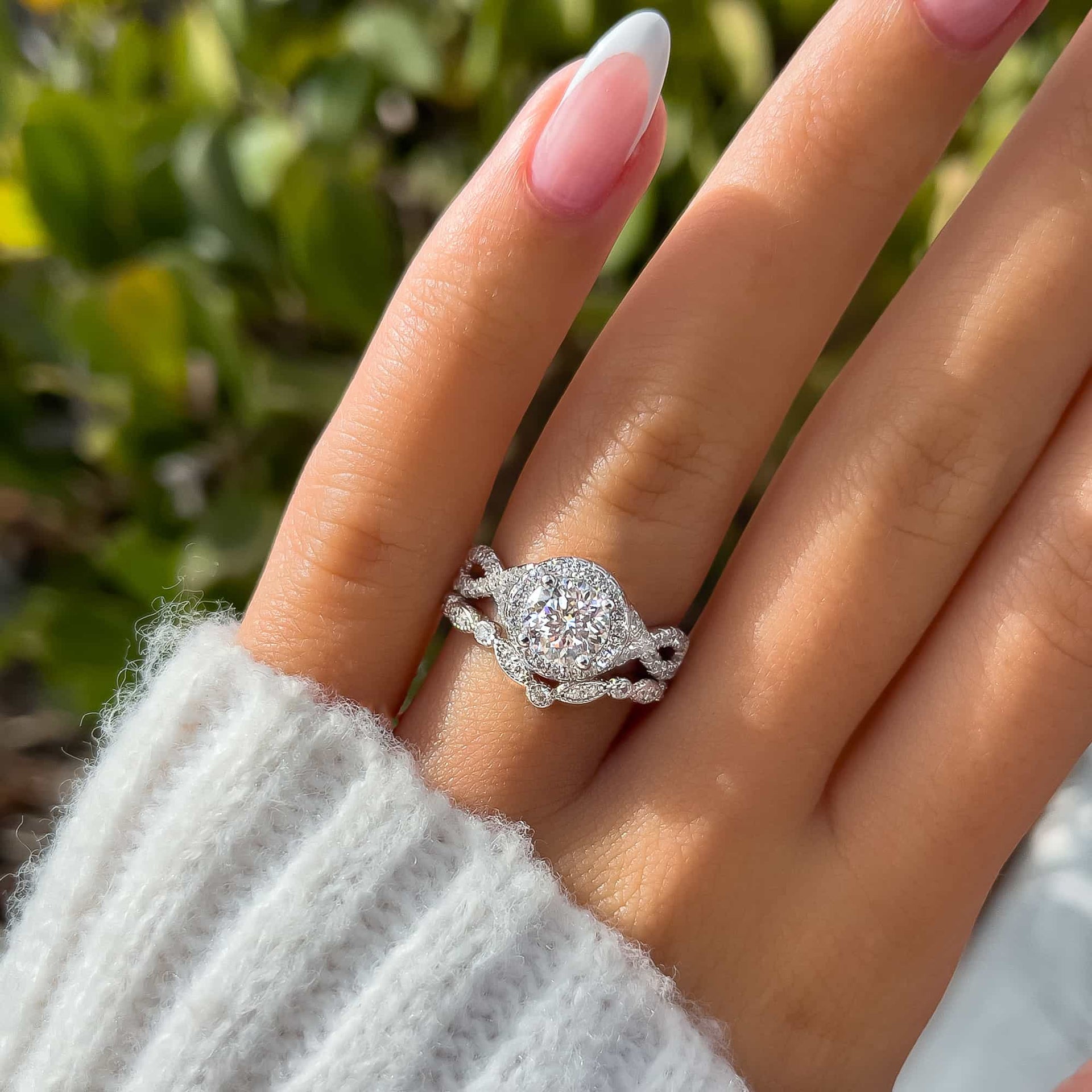 gorgeous silver 1.5 carat round cut vintage style engagement ring shown paired with a stunning silver chevron wedding band on model's french manicured nails with white sweater sleeve and greenery in the background