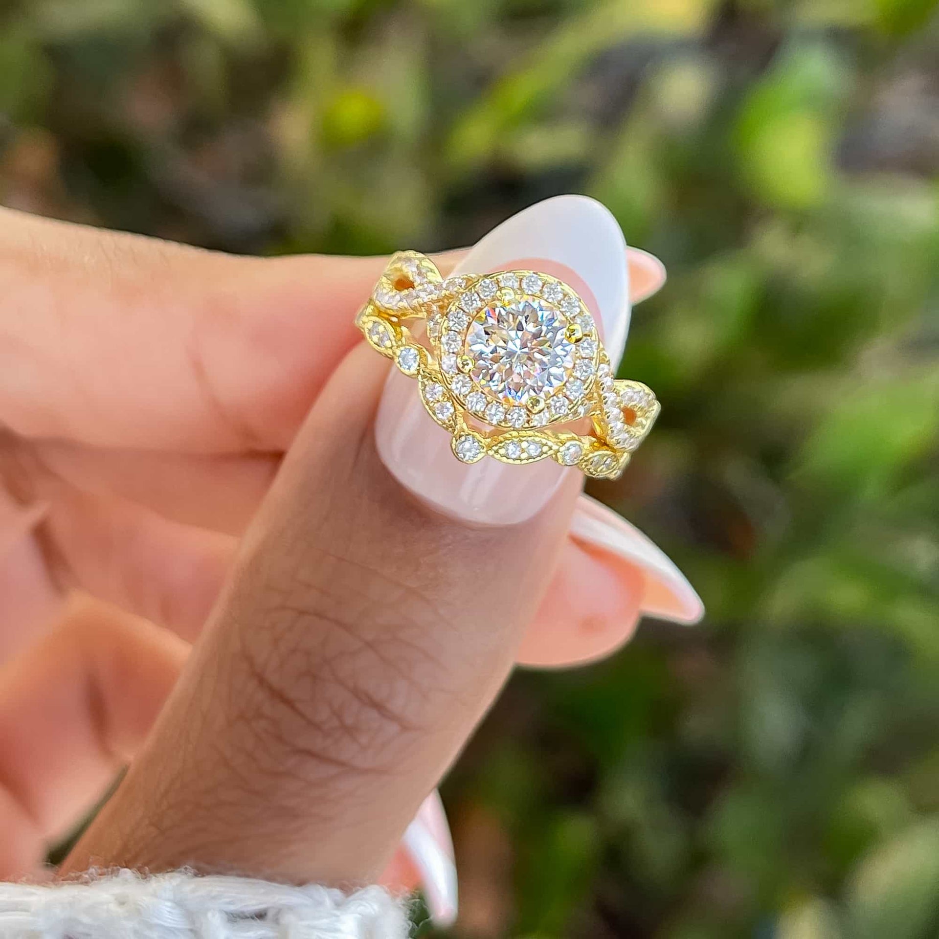 stunning gold chevron wedding band paired with a 1.5 carat round cut vintage style engagement ring shown on model's thumb with french tip and greenery in the background