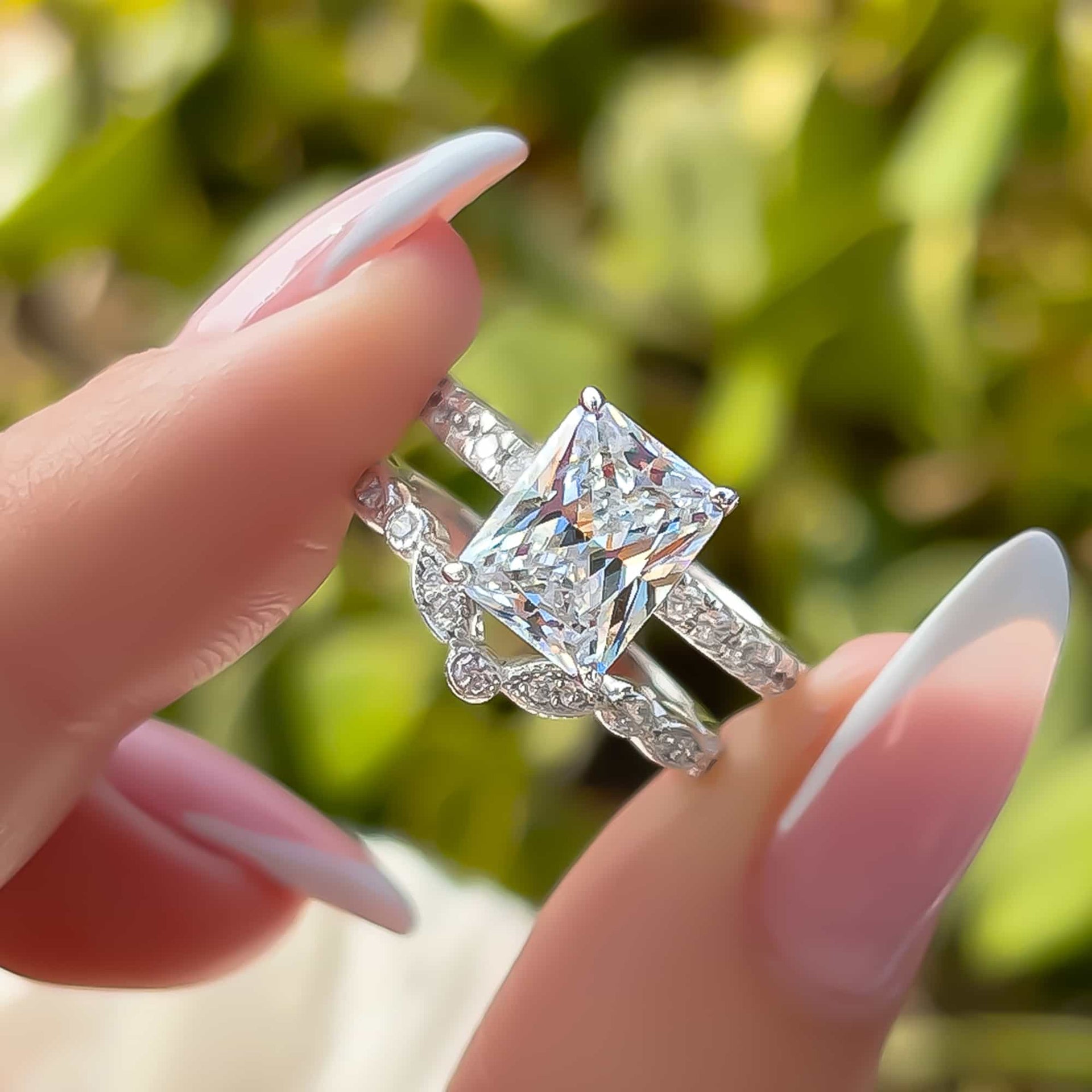 a stunning radiant cut silver engagement ring paired with a silver chevron wedding band being pinched between model's french manicured fingers with greenery in the background