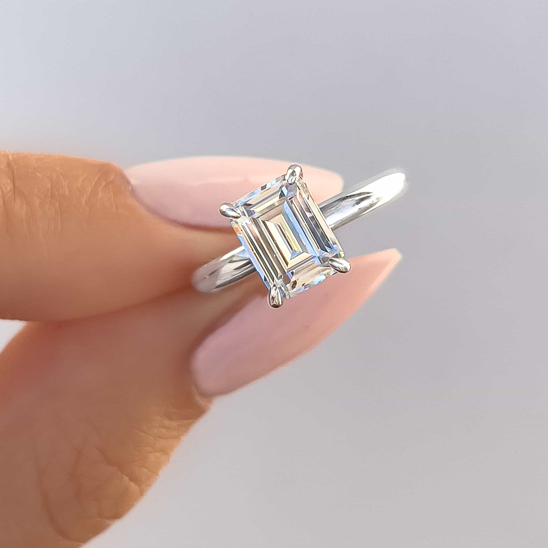 1.5 carat emerald cut silver solitaire engagement ring held by model with light pink nails