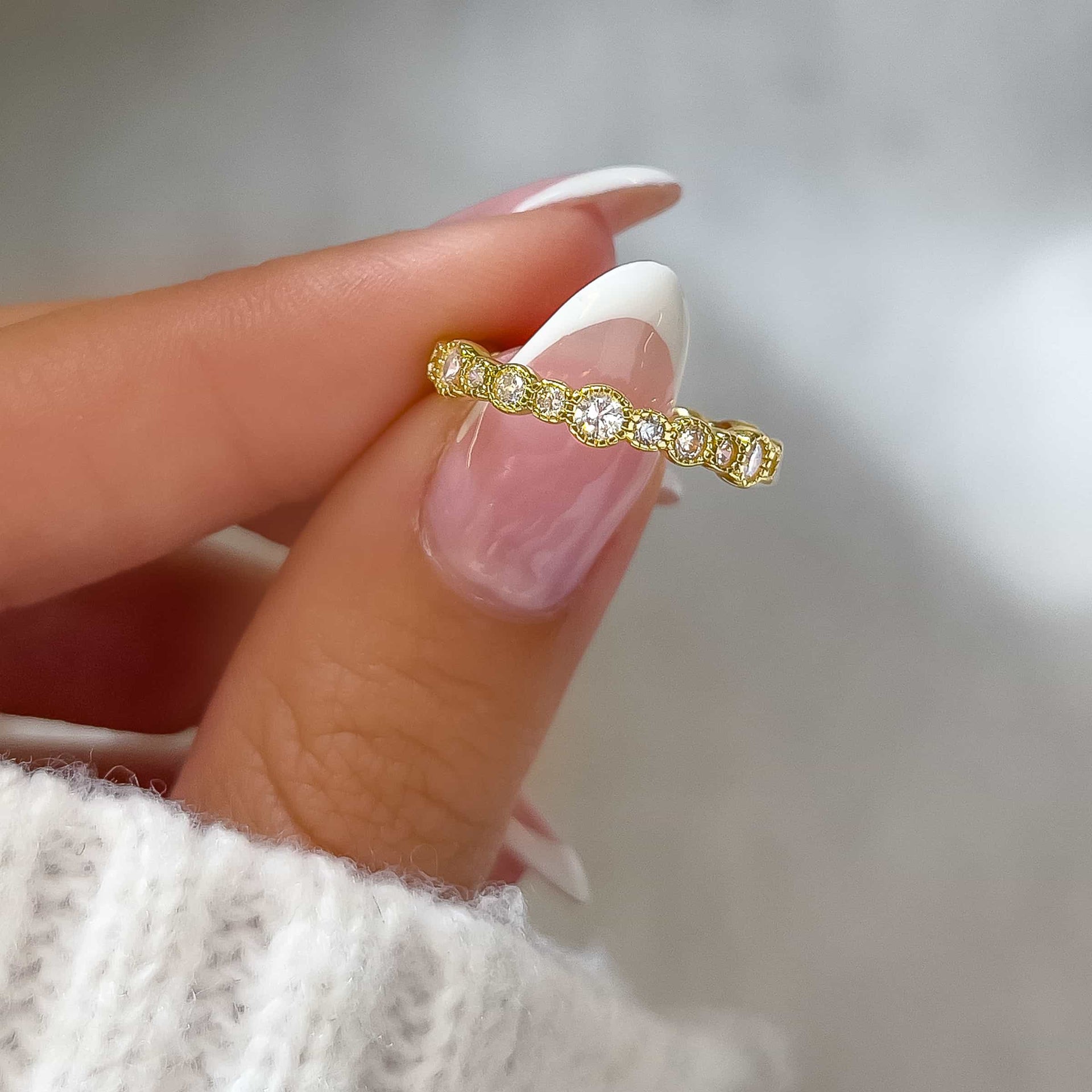 shiny unique multi-stone wedding band held by model with french tip nails and white sweater