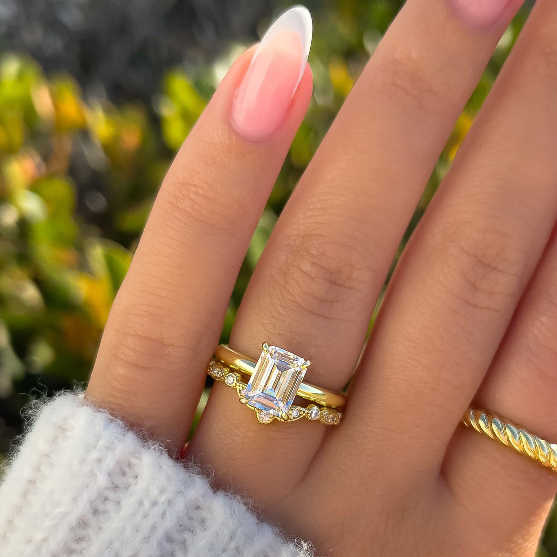 Gold wedding ring set made up of an emerald cut solitaire engagement ring and chevron half eternity wedding band on model with french tip nails