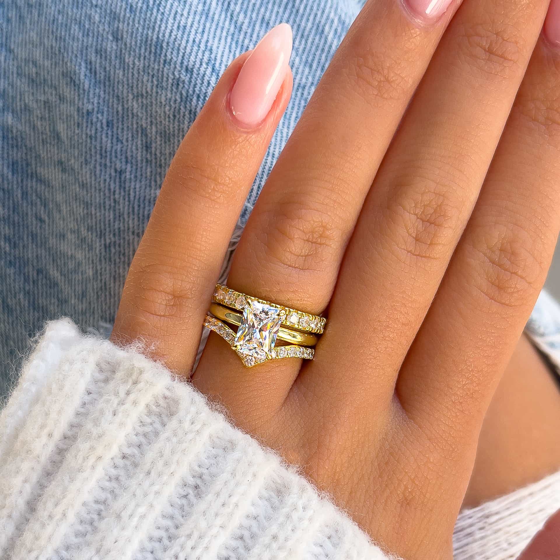 stunning gold 1.5 carat radiant cut solitaire engagement ring paired with two gold wedding bands on model with neutral nails and white sweater