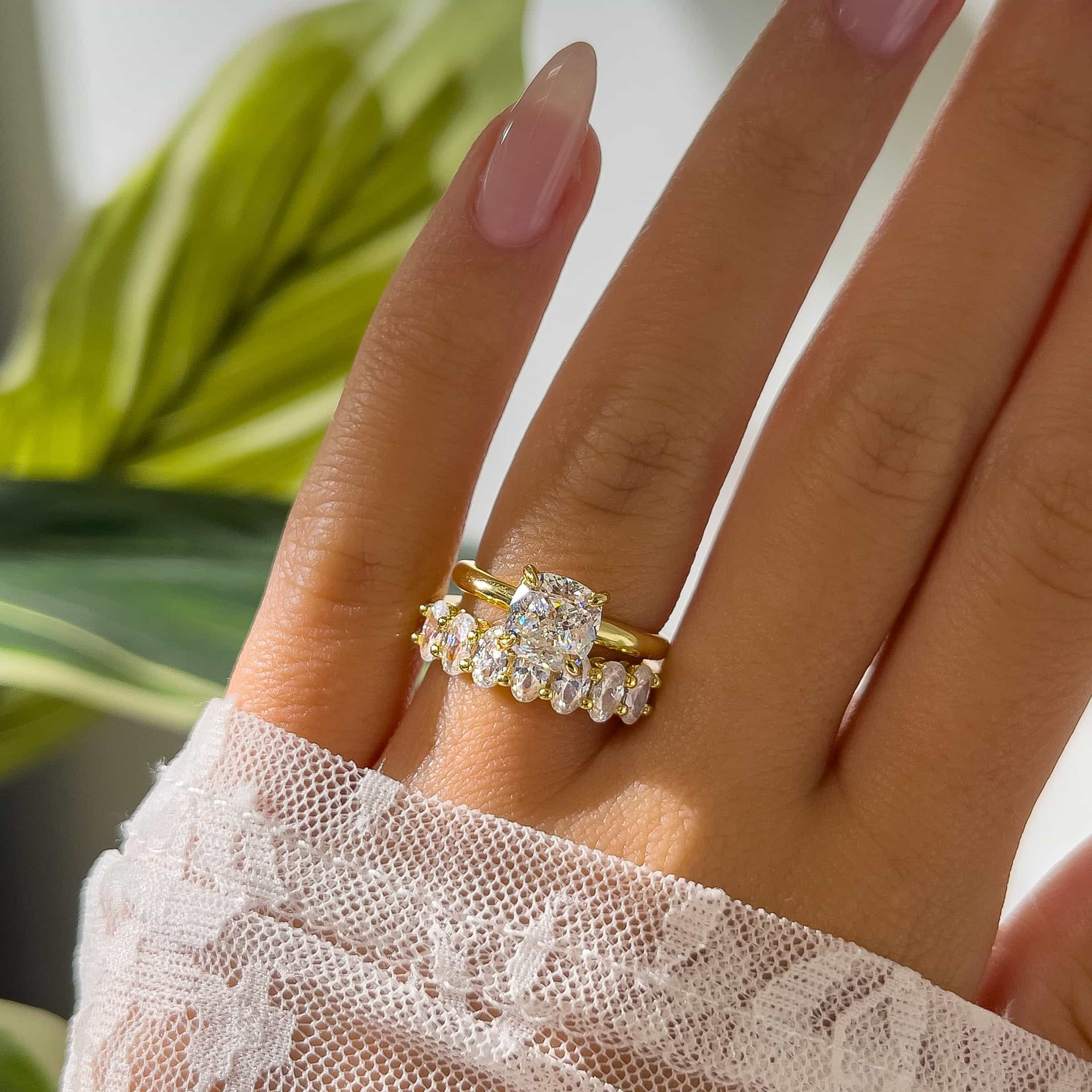 gold oval cut half eternity wedding band paired with gold cushion cut engagement ring with white lace sleeve and greenery in the background