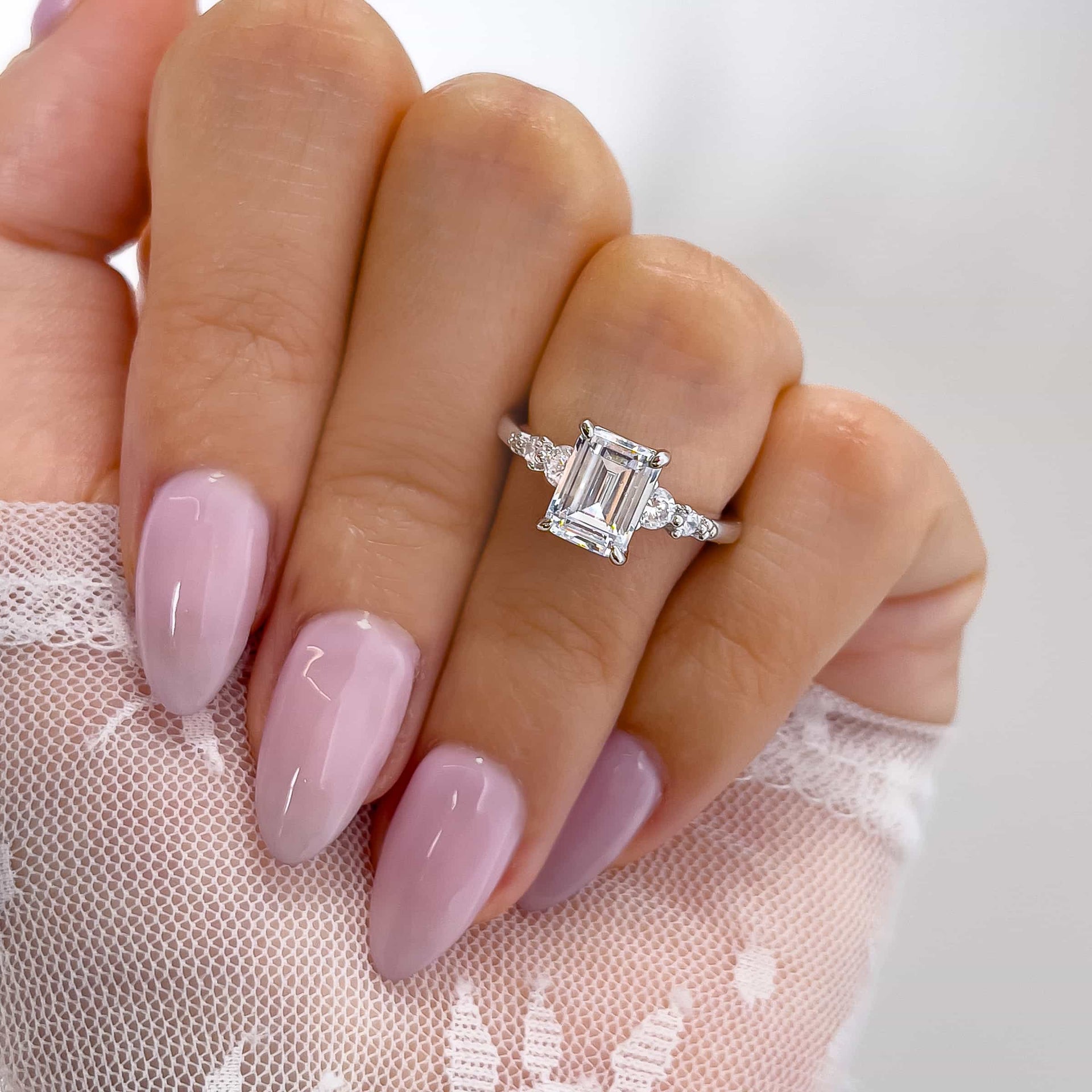 classic 1.5 carat emerald cut engagement ring with round accent stones on both sides on model with lace top and light pink nails