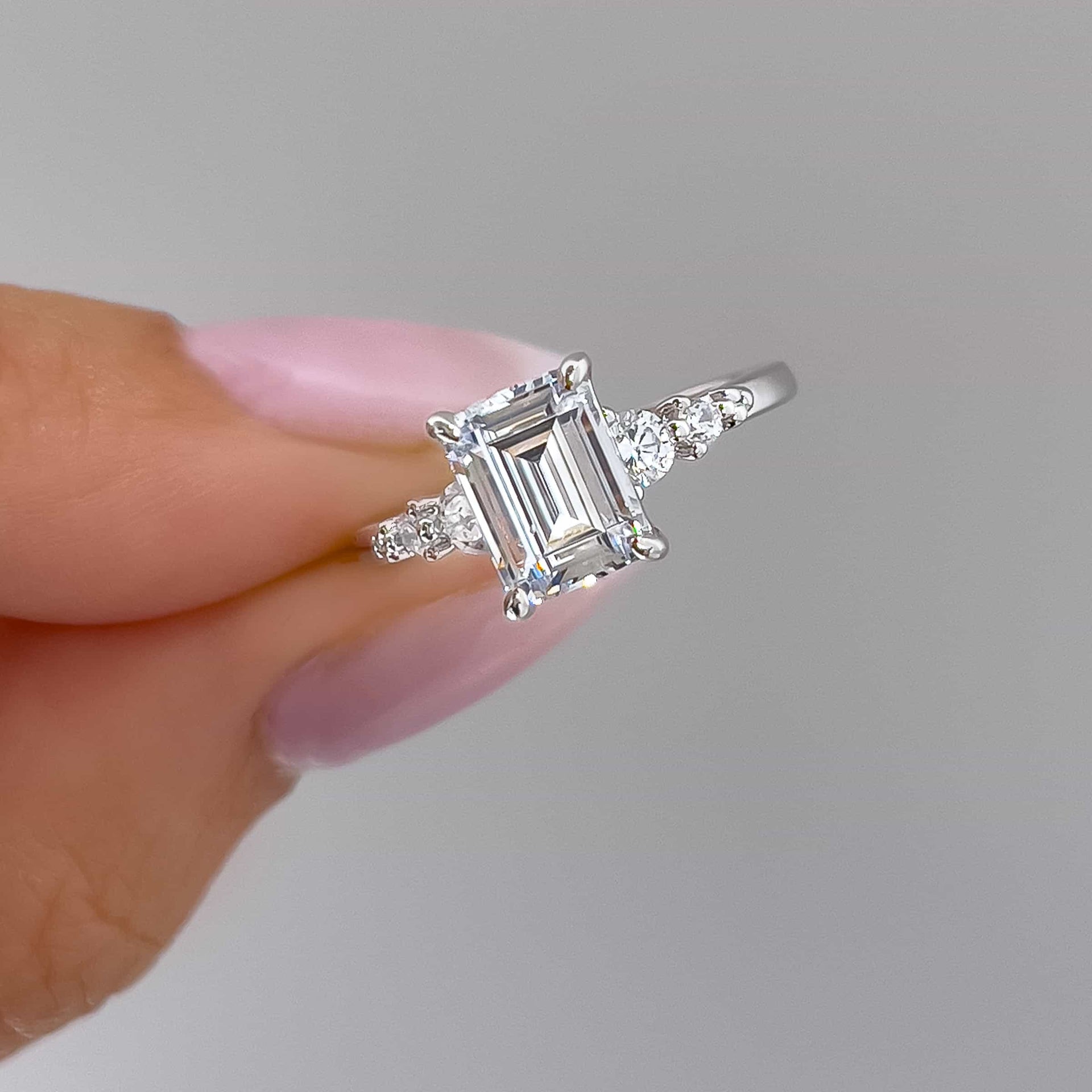 close up of silver emerald cut engagement ring held by model with light pink nais