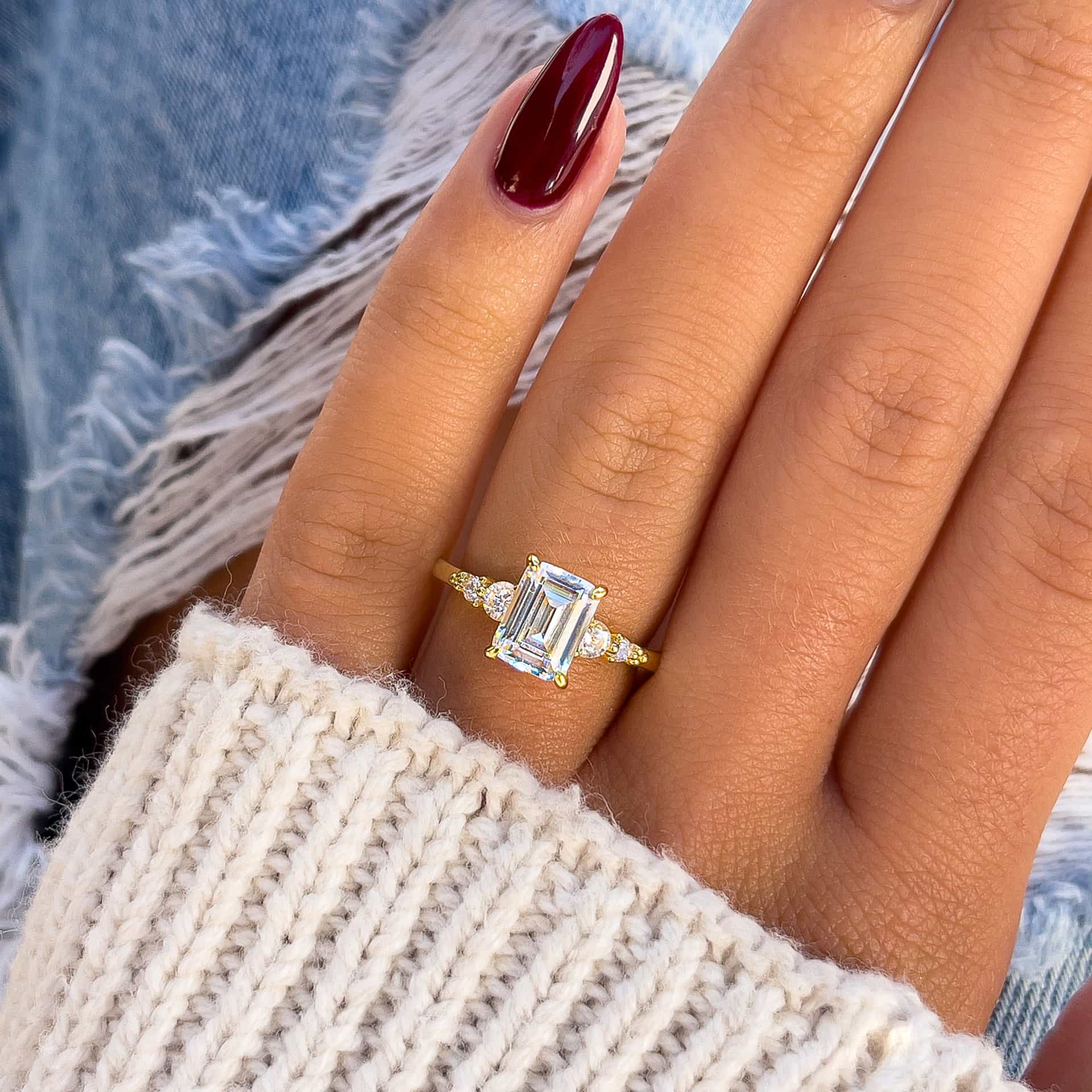 simple gold 1.5 emerald cut engagement ring on model with red nails and cream sweater