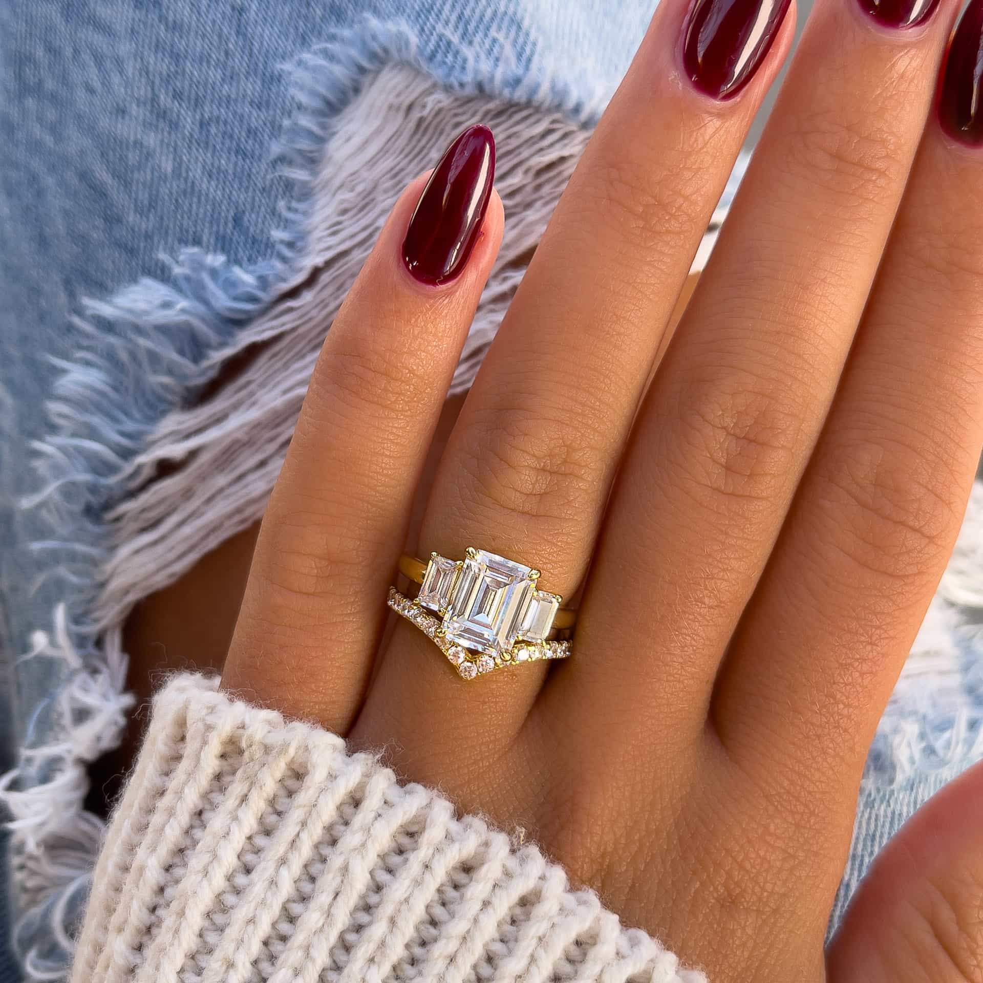 stunning and unique wedding ring set made up of a gold 1.5 carat emerald cut engagement ring and half eternity chevron band on model with burgundy nails, neutral colored sweater, and ripped jeans