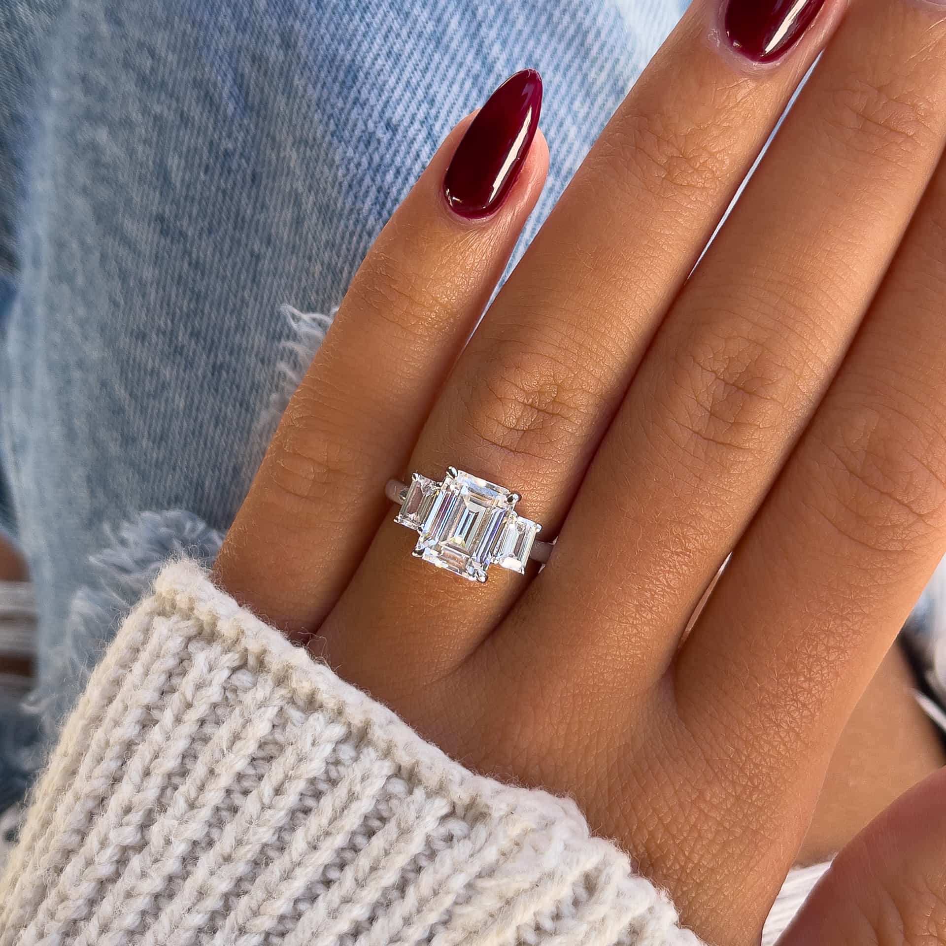 stunning silver emerald cut trio engagement ring shown on model's hand with deep red nails and a cream sweater