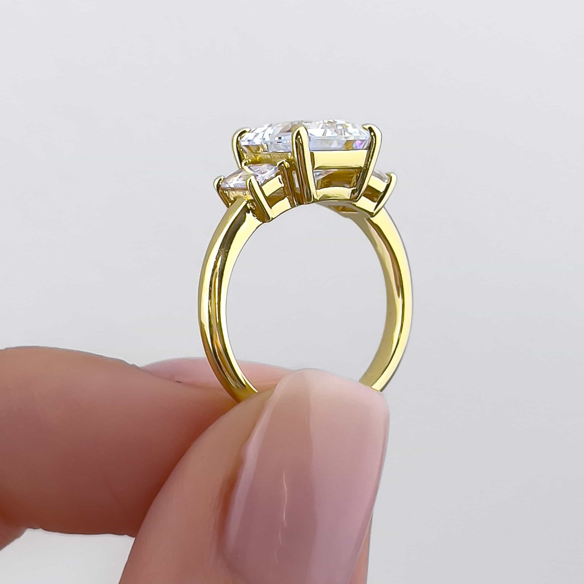 setting shot of a gold engagement ring featuring a 1.5 carat emerald cut center stone and two complementary .5 carat emerald cut stones on both sides, held by model with neutral nails