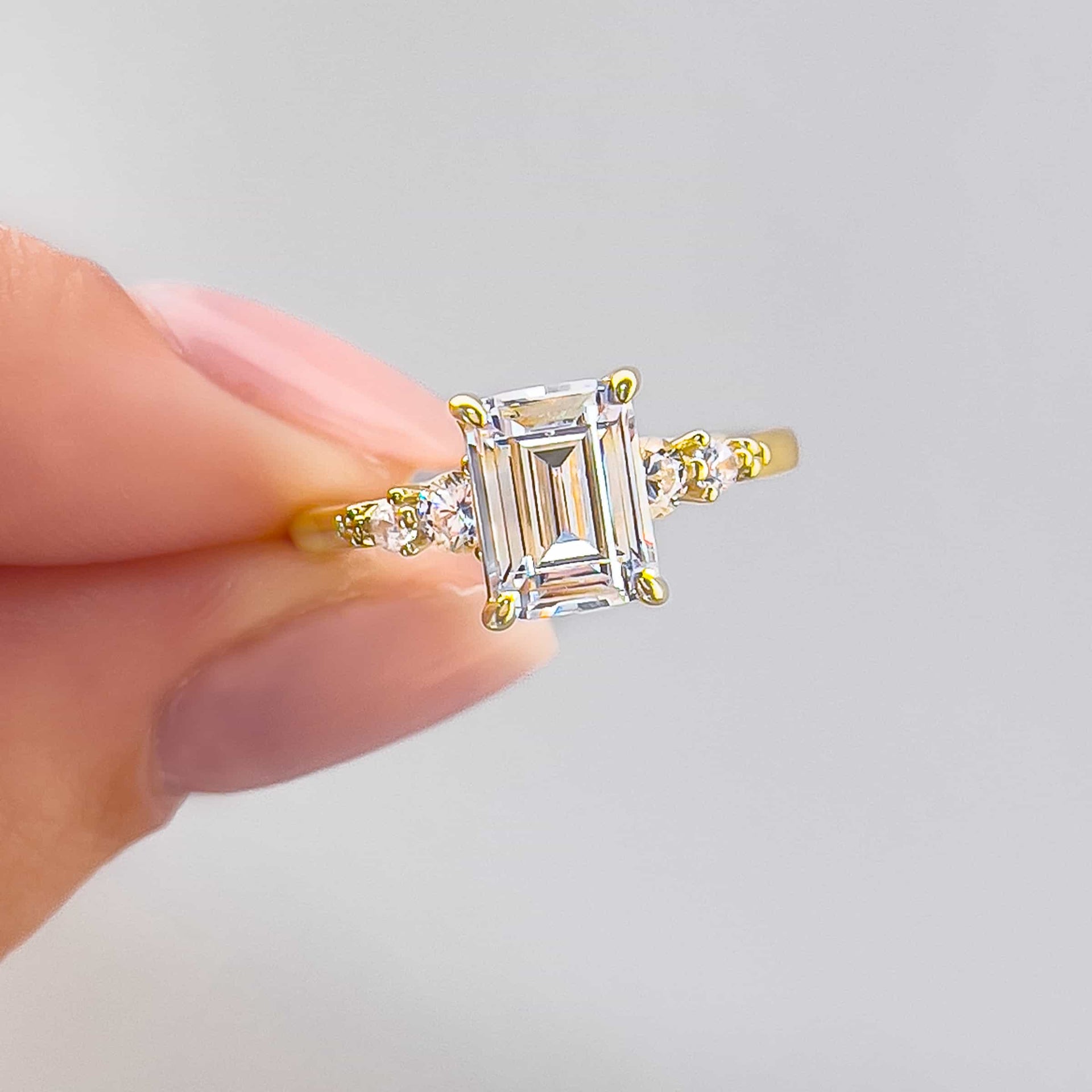 close up photo of gold 1.5 carat emerald cut engagement ring with two round cut side stones on each side held by model with neutral nails