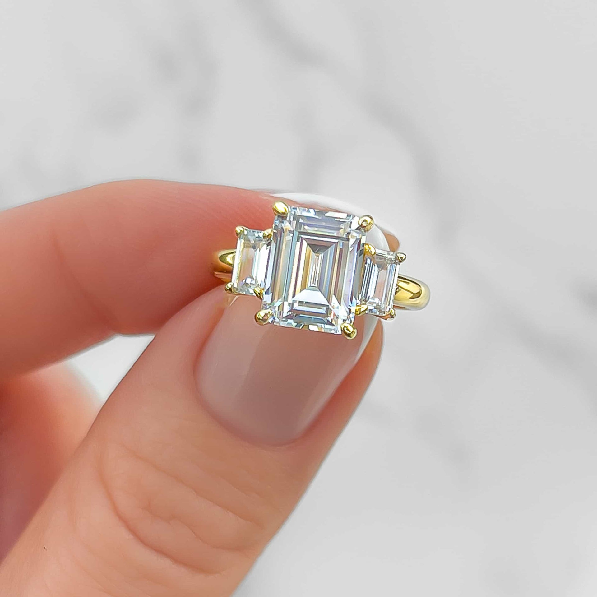 close up of gold, trinity style emerald cut engagement ring held by model with french tip nails