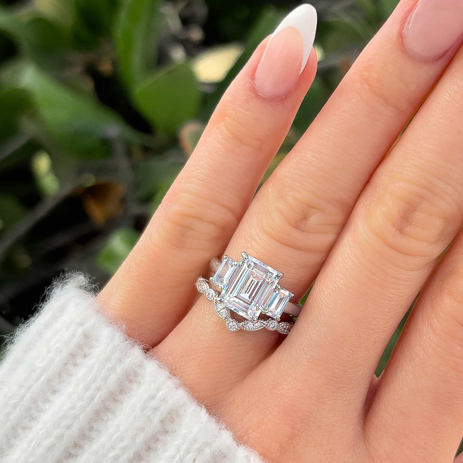 stunning silver emerald cut trio engagement ring paired with a vintage chevron style band on model's hand with french tip nails and white sweater