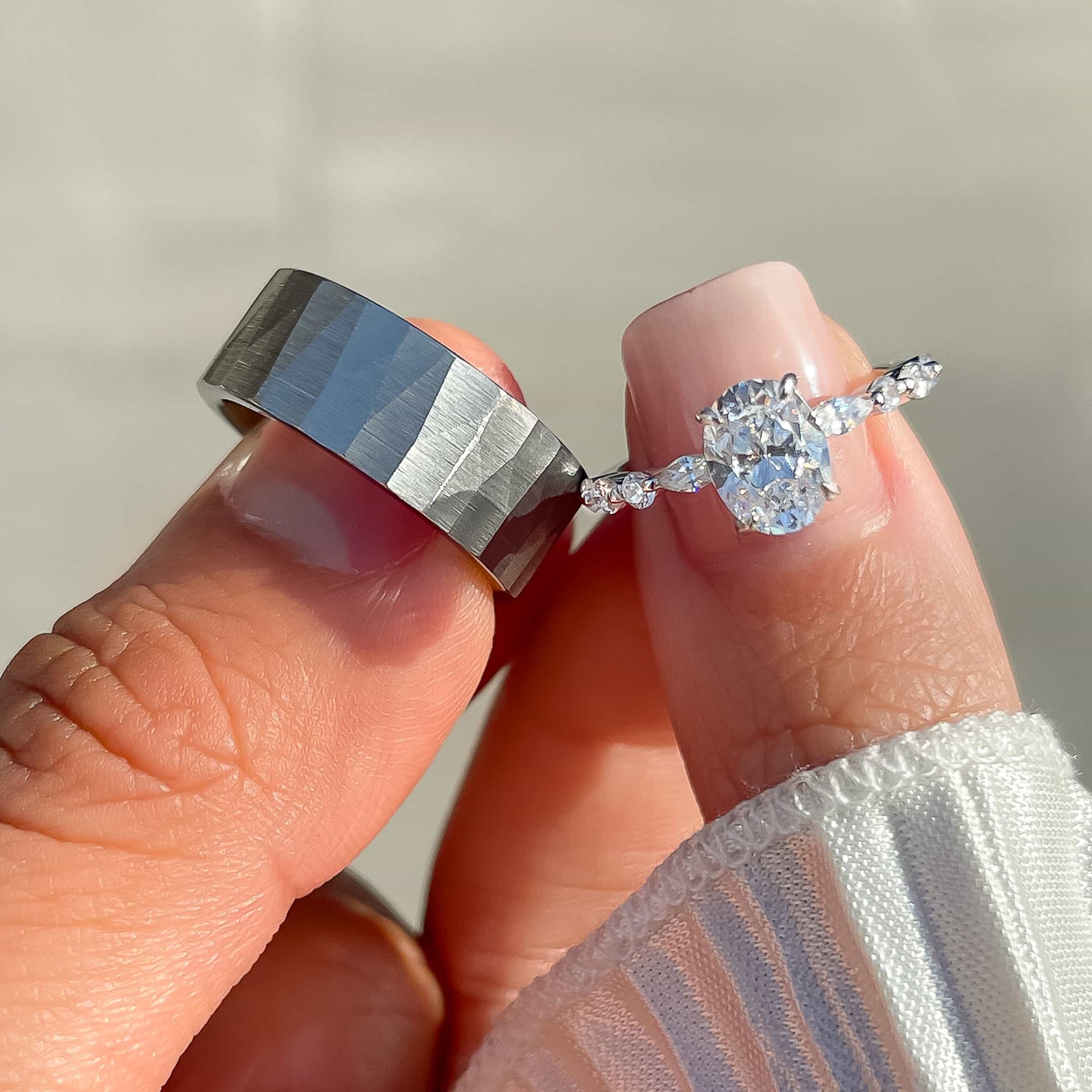 unique texture silver men's wedding band shown with silver oval cut engagement ring. each ring is held by a male and female model.