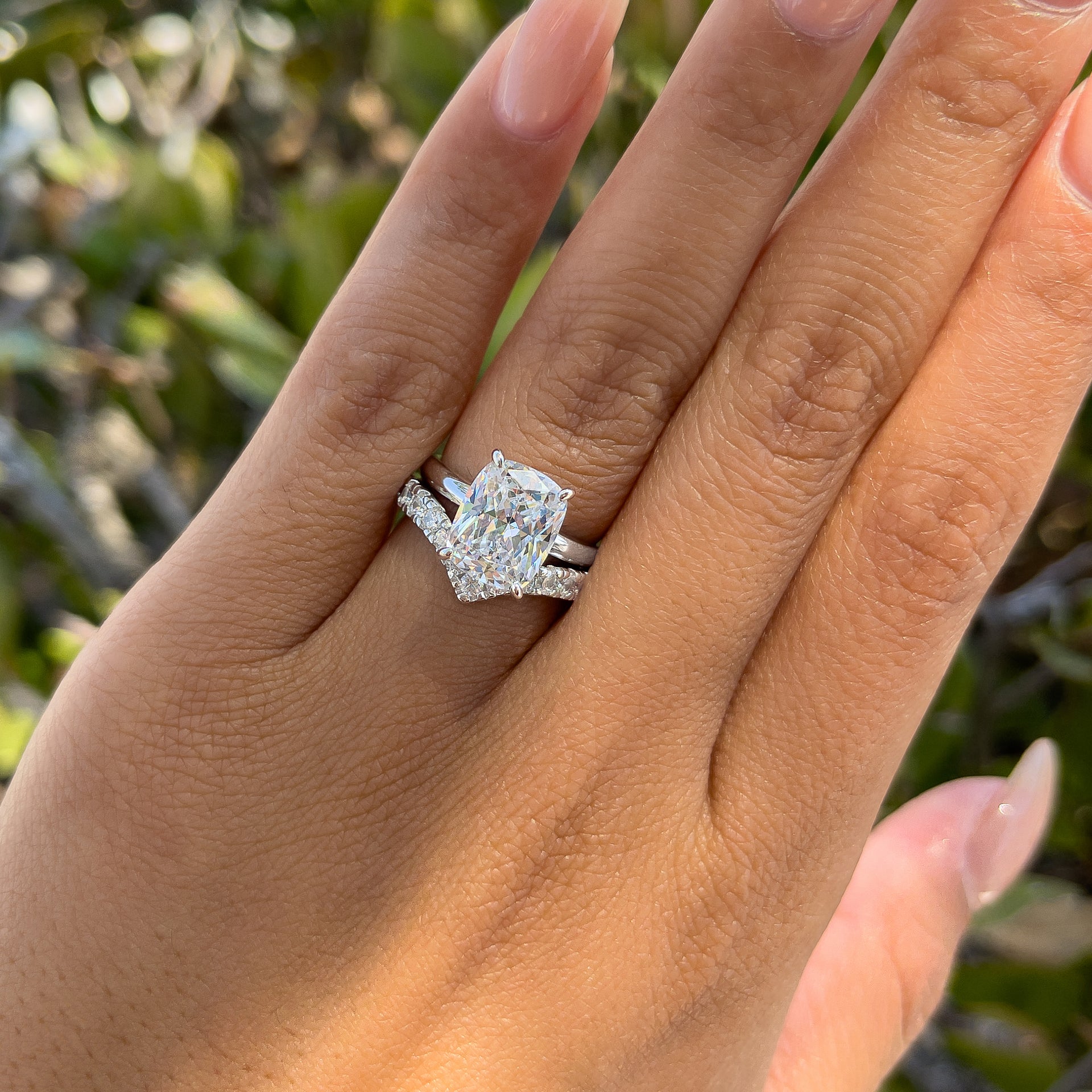 Stunning silver wedding set made up of a 4 carat elongated cushion cut solitaire and silver chevron wedding band, worn by model with neutral nails in front of greenery