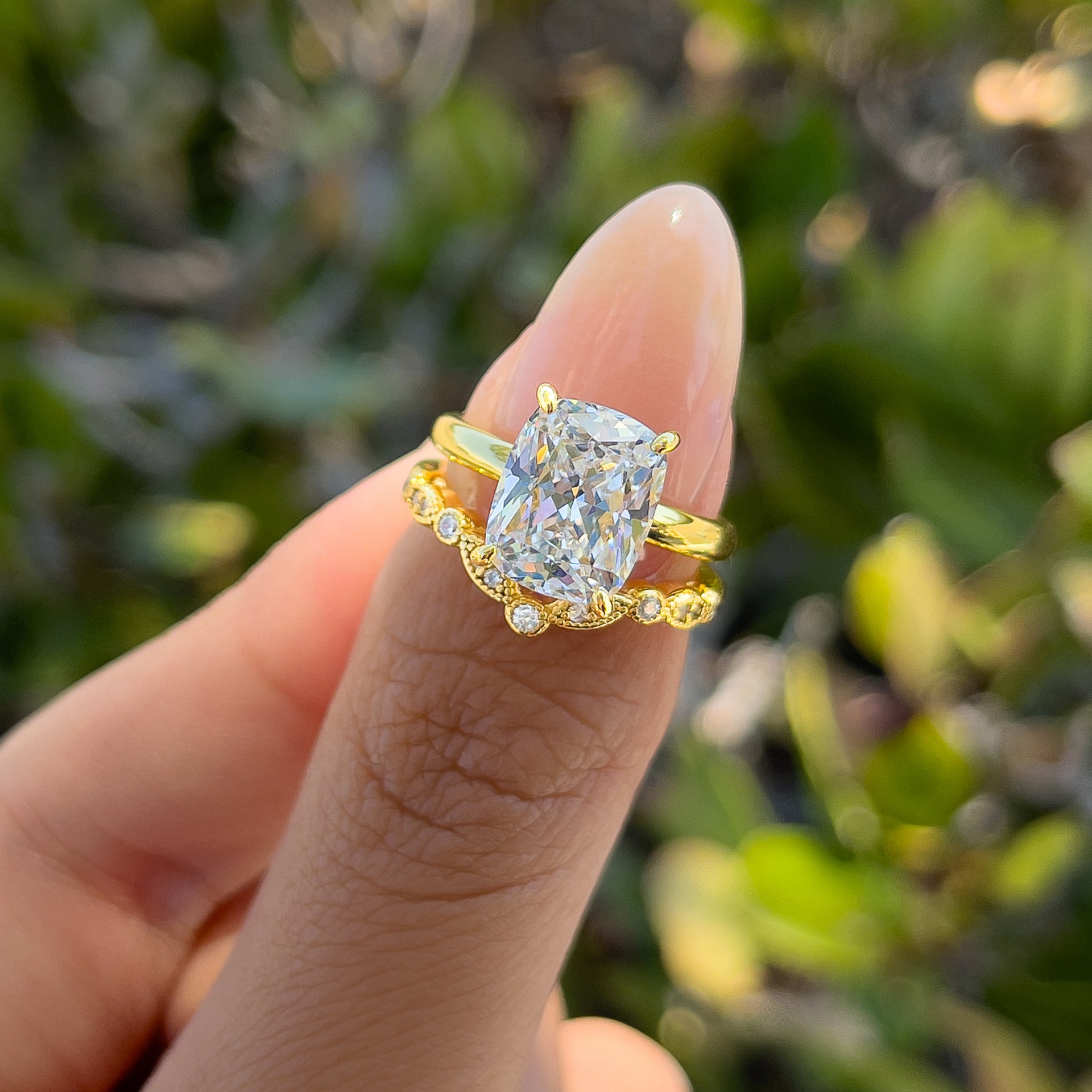 Stunning gold wedding set made up of a 4 carat elongated cushion cut solitaire and vintage design gold chevron band, held by model with neutral nails in front of greenery