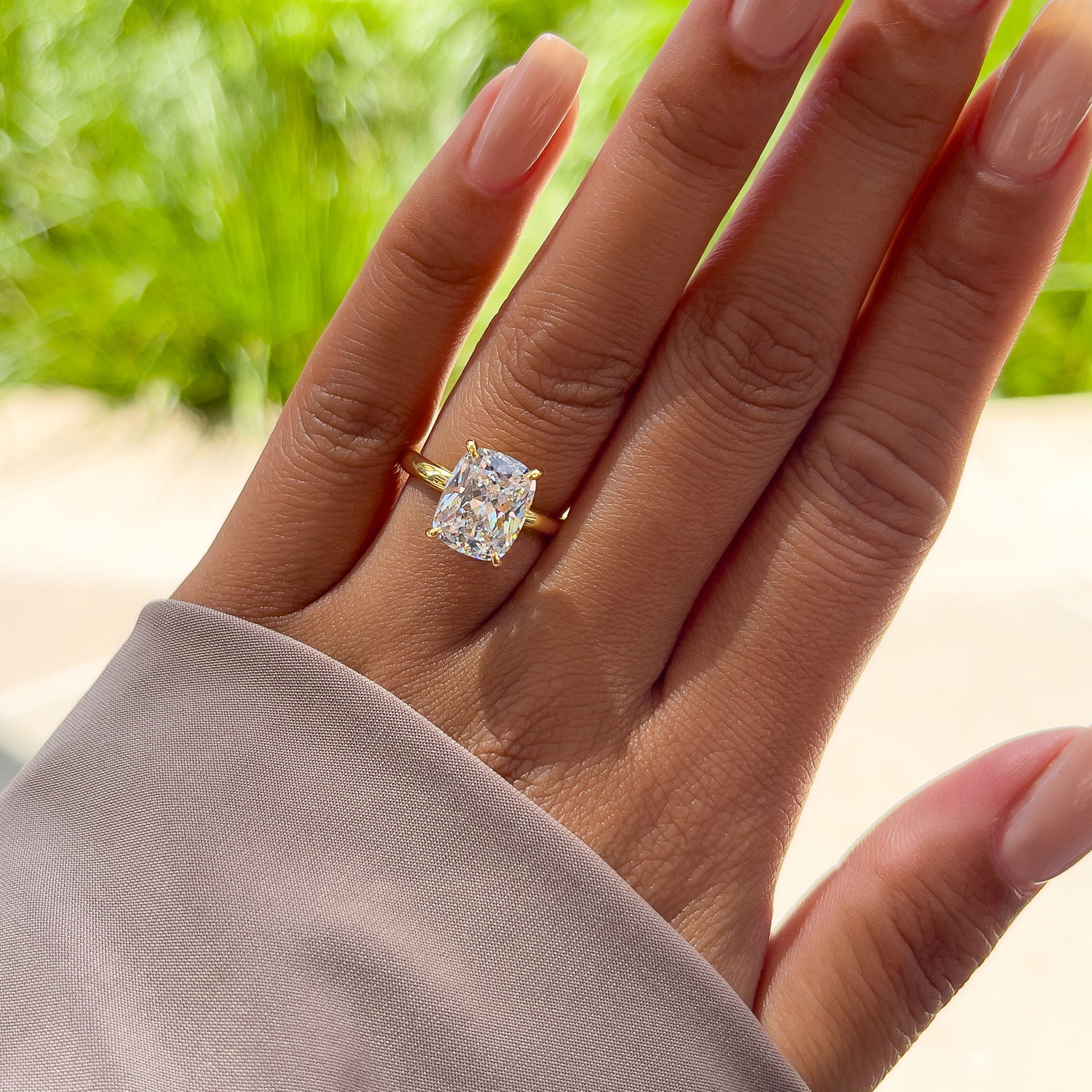 Simple gold elongated cushion cut solitaire engagement ring on model with neutral nails in front of greenery