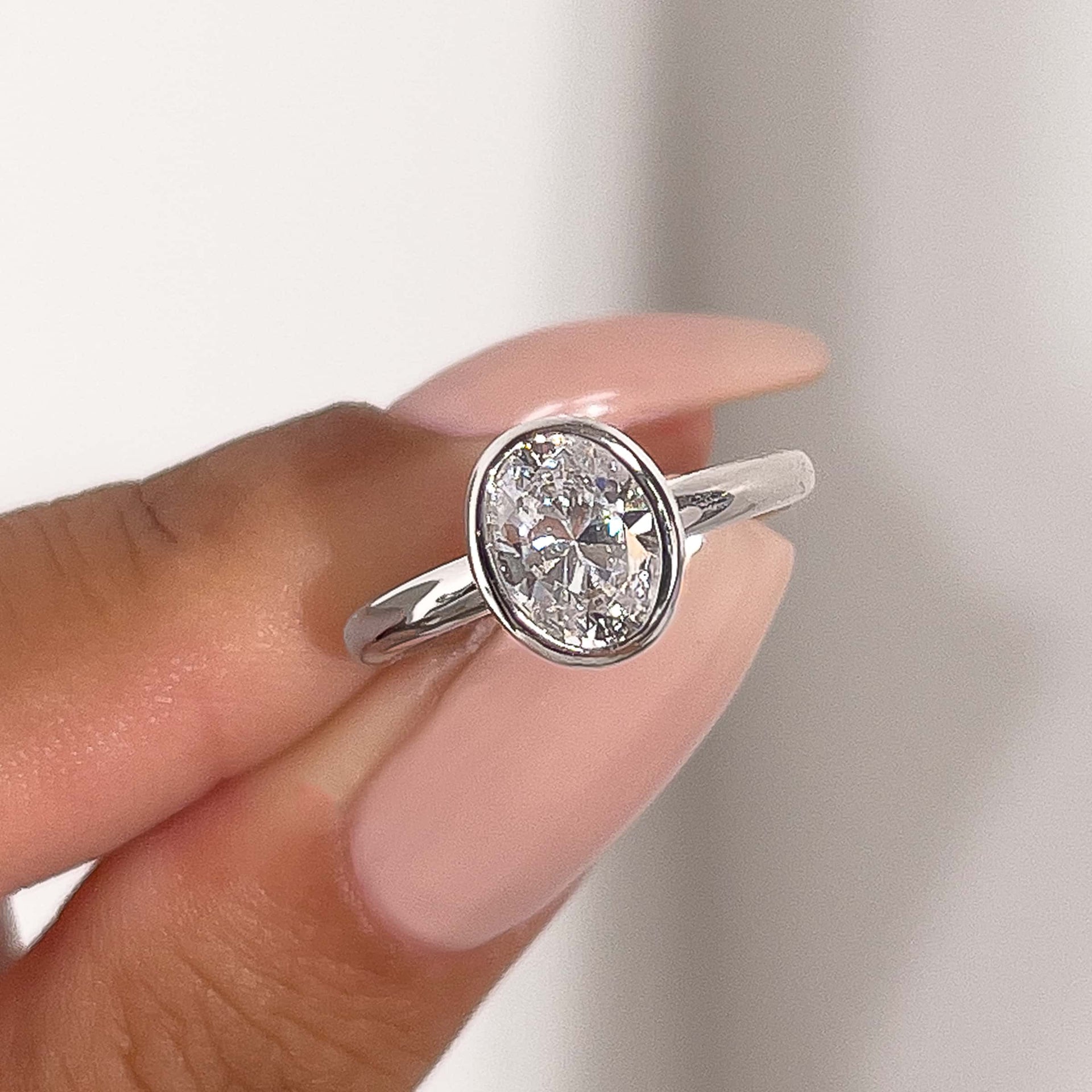 a beautiful silver oval cut bezel silver engagement ring pinched between a woman's fingers with pink manicured nails 