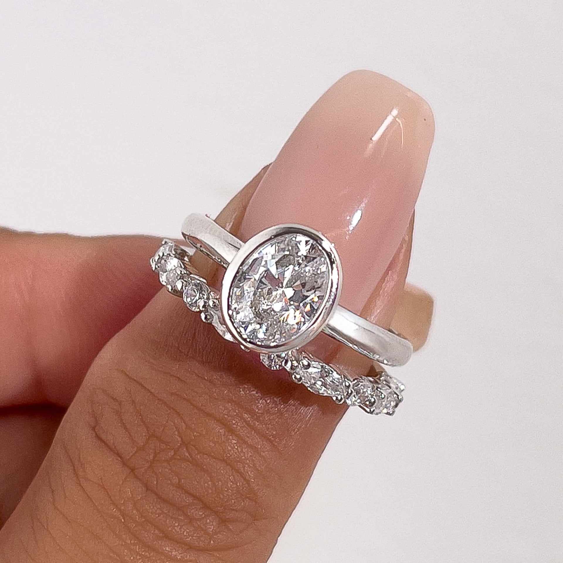 a close-up shot of a silver bezel oval cut ring with a pave silver wedding band on model's thumb with pink manicured nails