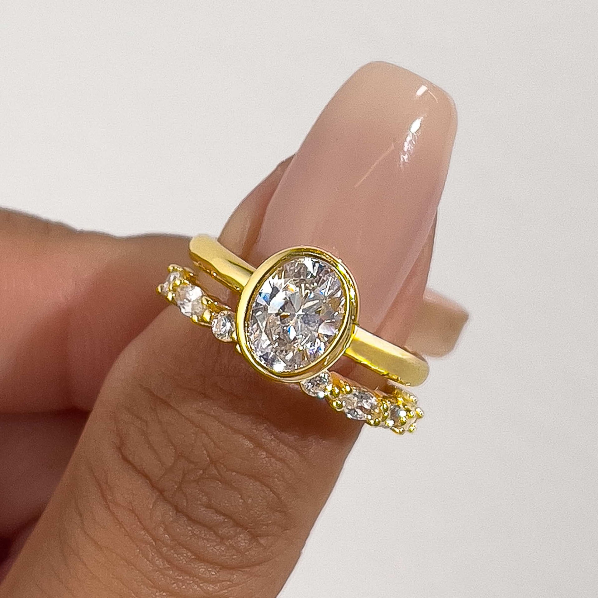 a close-up shot of a gold bezel oval cut ring with a pave gold wedding band on model's thumb with pink manicured nails