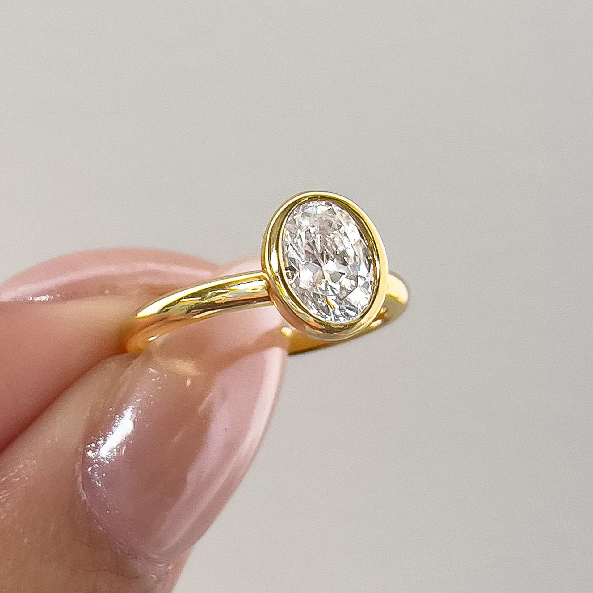 a gorgeous slightly angled shot of an oval cut gold bezel engagement ring pinched between models fingers with light pink shimmering nails