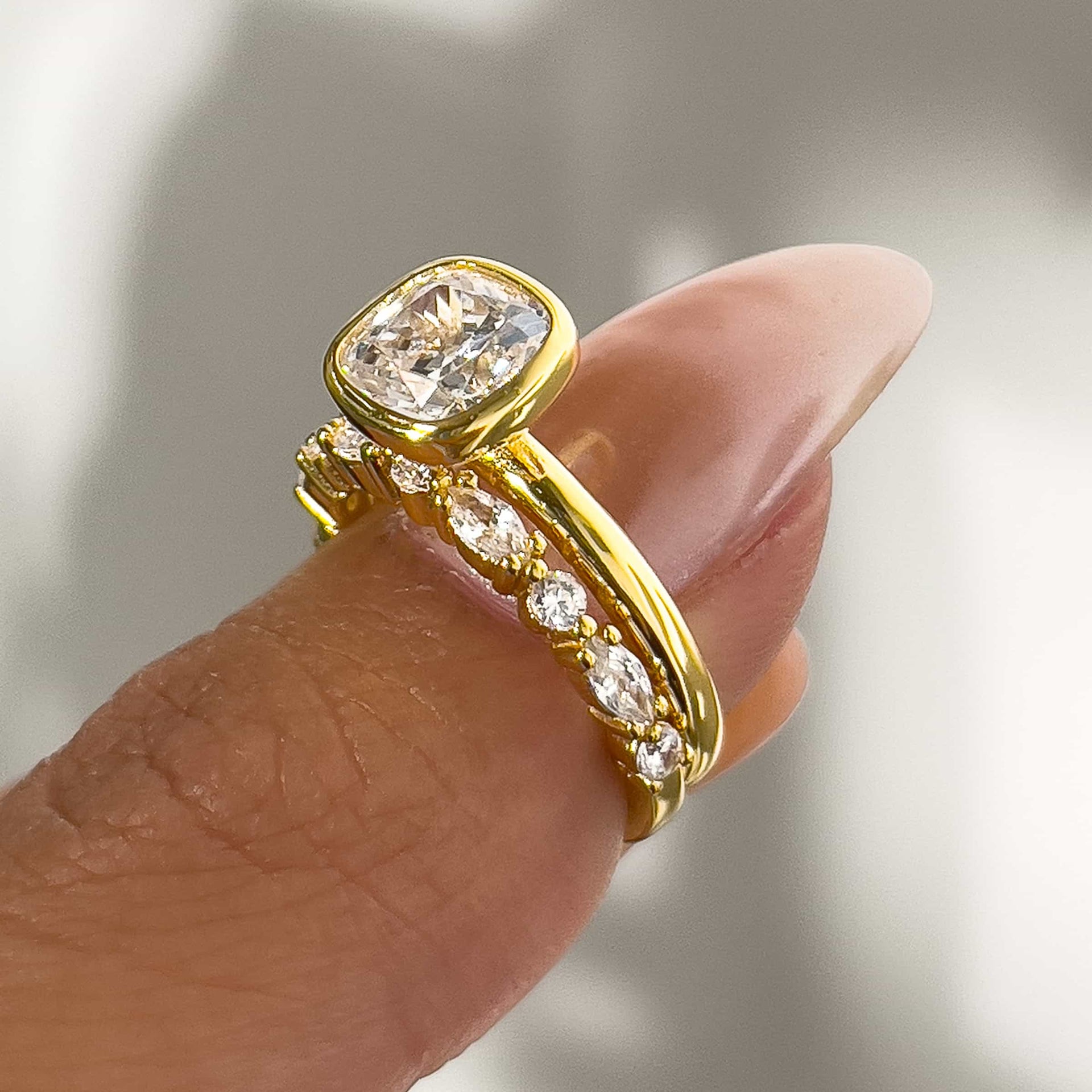 a beautiful gold cushion-cut bezel engagement ring with a mix-cut pave wedding band on model's thumb with white background