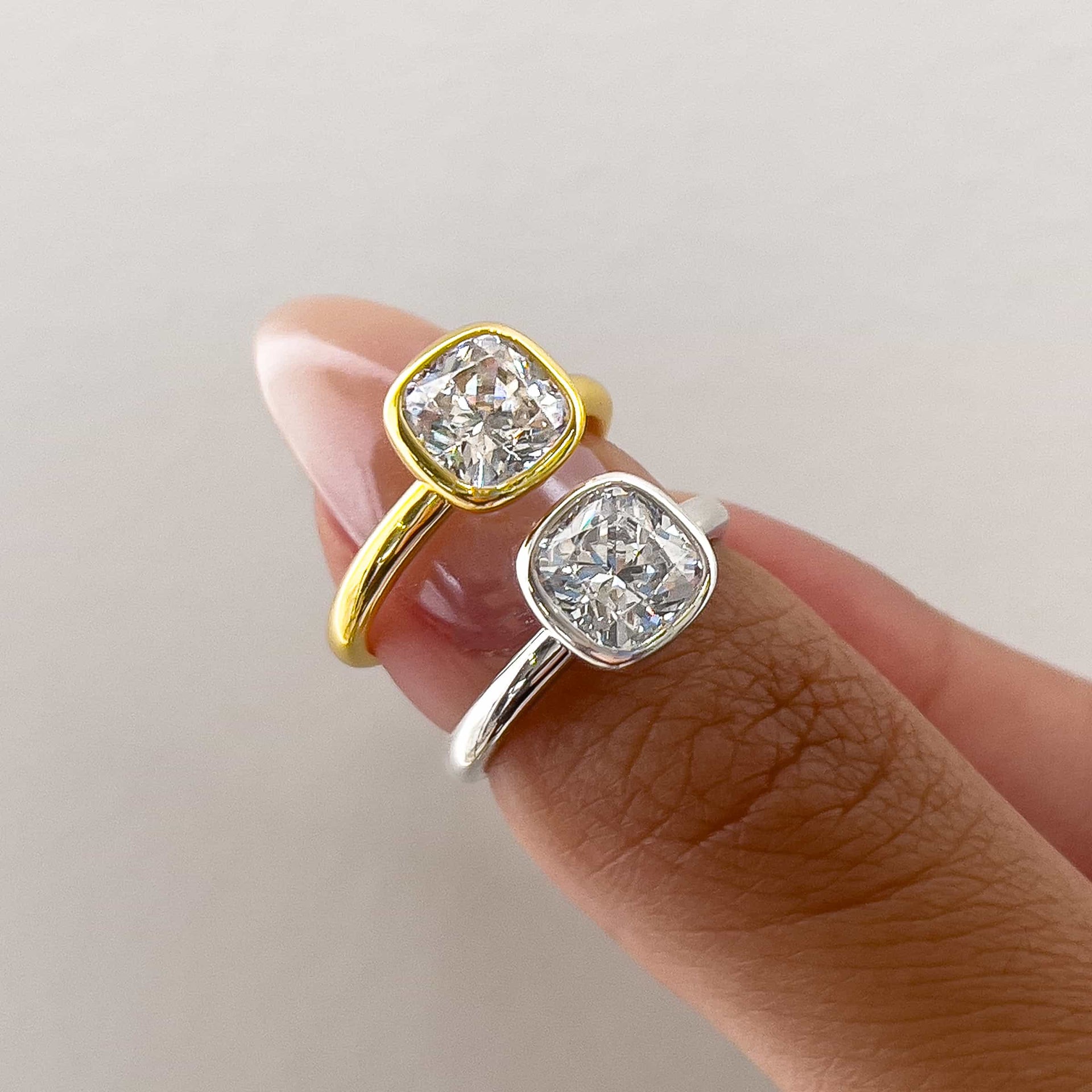 two beautiful cushion cut engagement rings in gold and silver on model's thumb with manicured pink nails and a white background