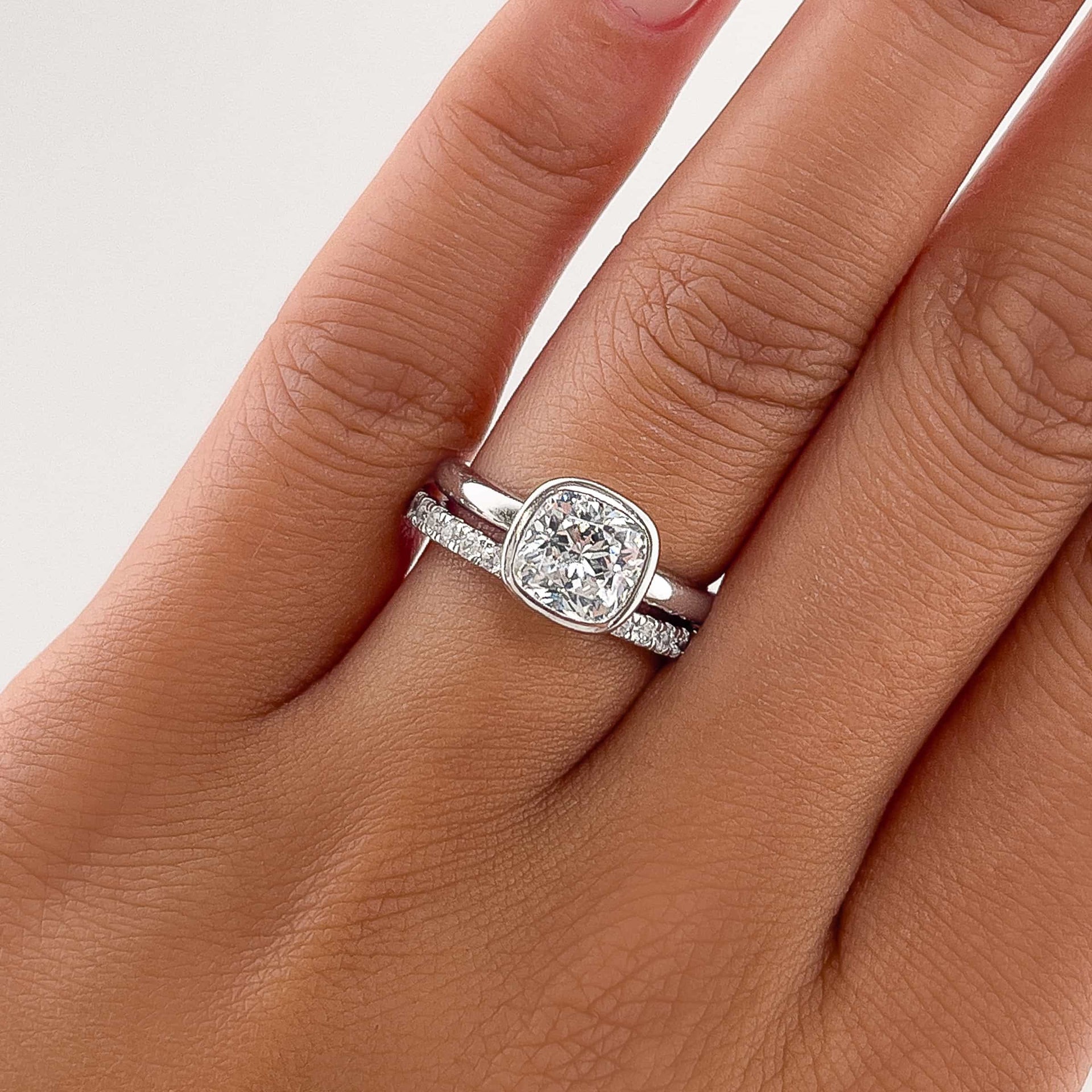 a beautiful silver bezel cushion cut engagement ring on model's ring finger with a silver eternity wedding band paired and a white backround