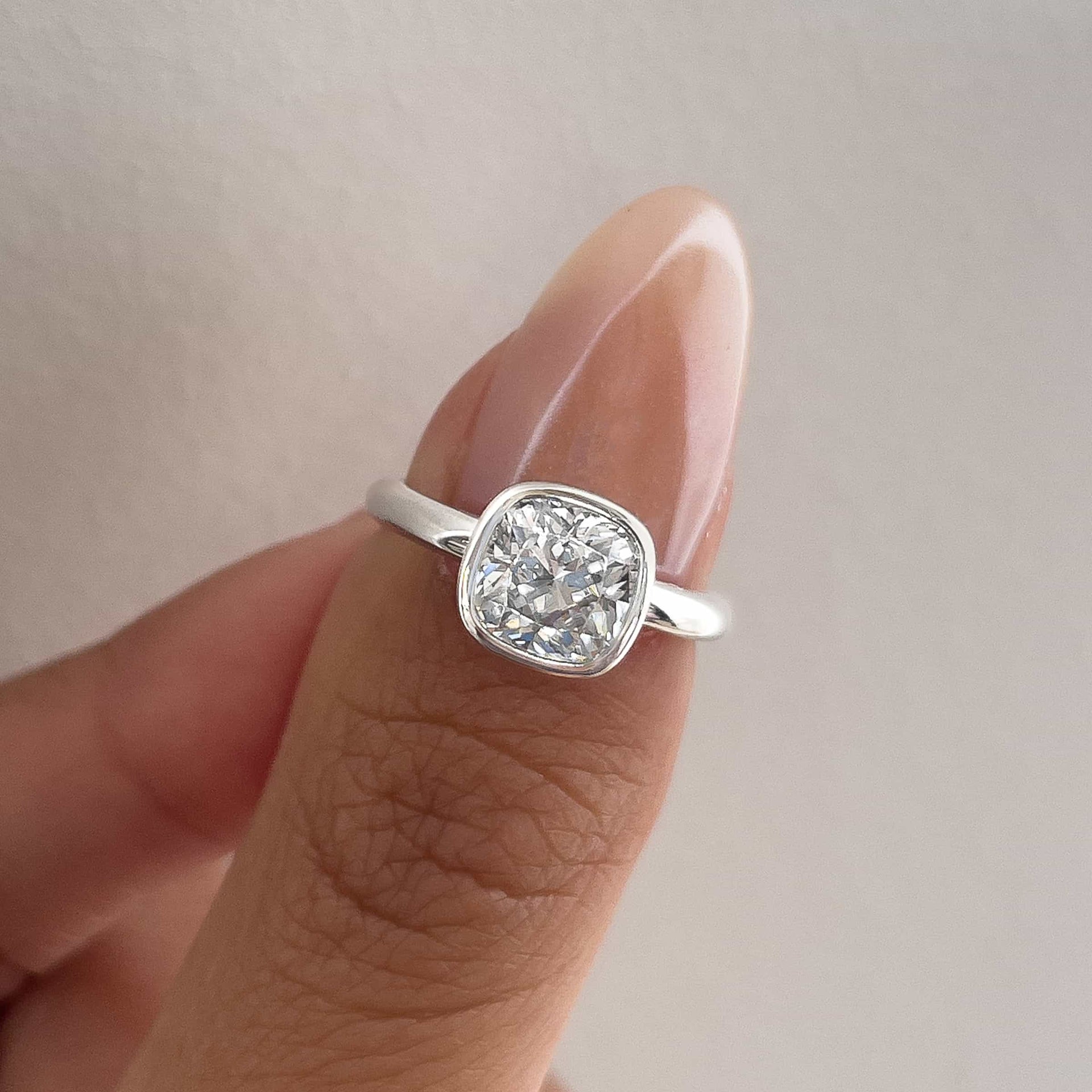 a stunning silver cushion cut bezel engagement ring on model's thumb with pink muted nails and a white background