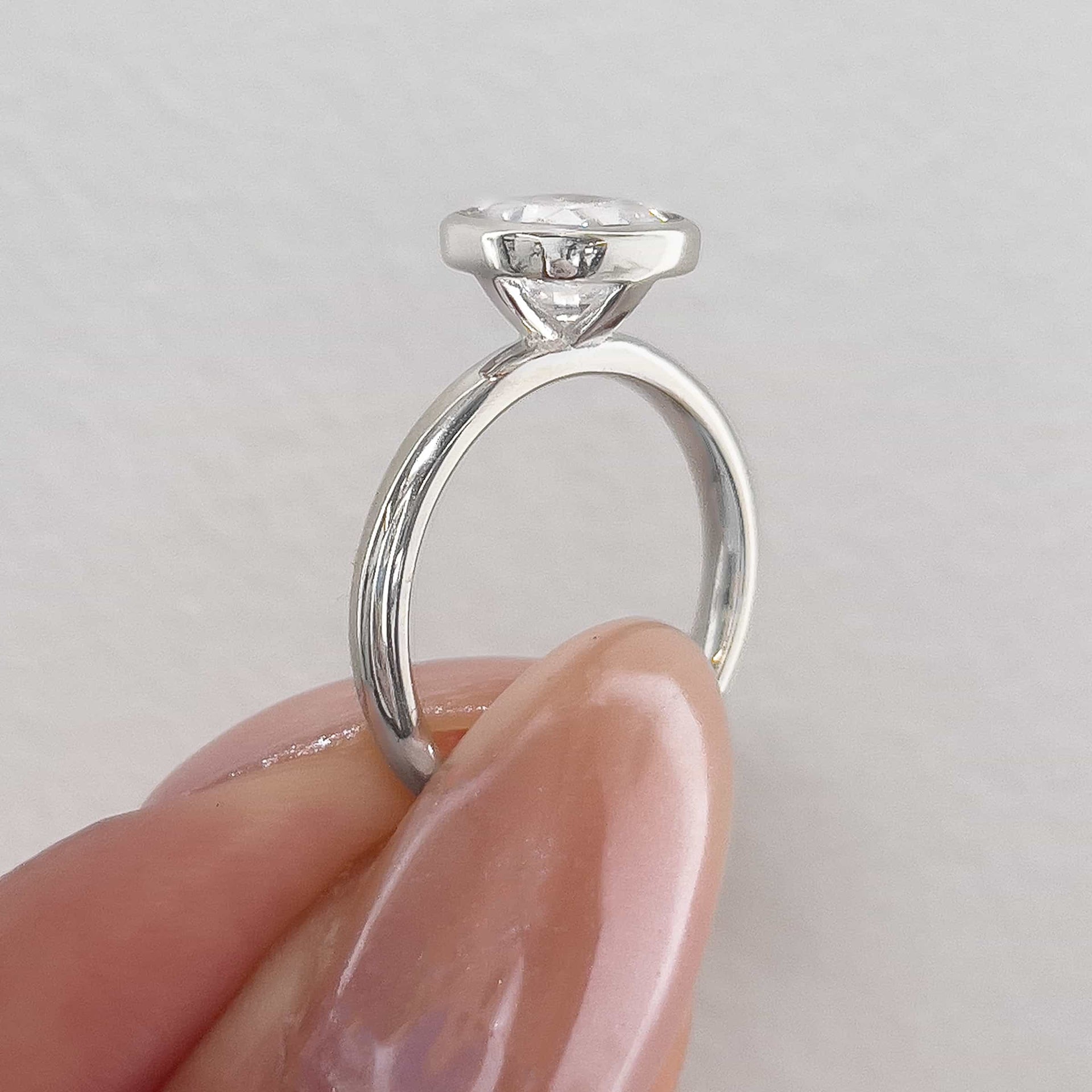 a setting shot of a stunning silver cushion cut bezel engagement ring pinched between model's thumb and pointer finger with white background