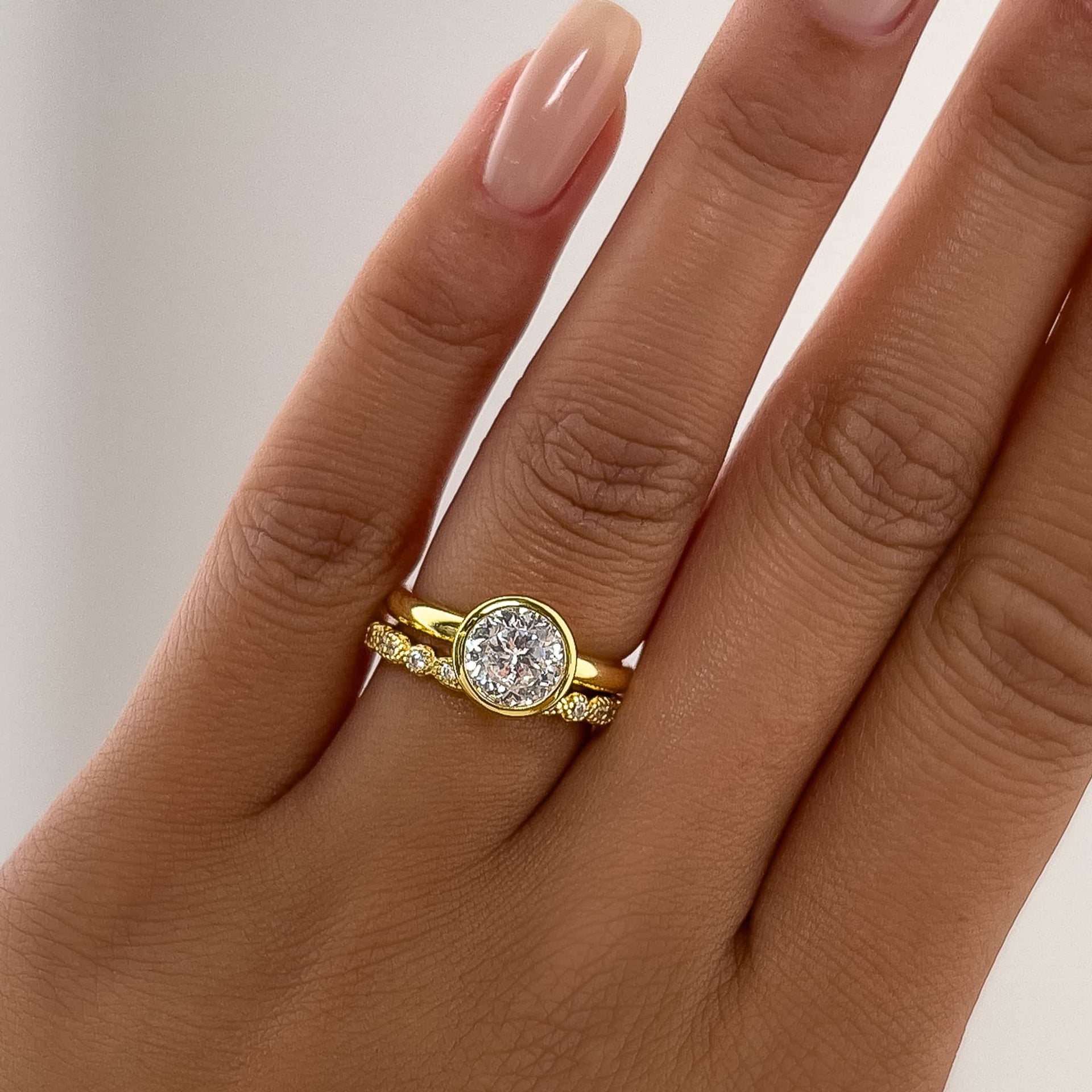 woman's hand with a gold 1.25 carat round cut bezel engagement ring and a pave wedding band on ring finger with neutral tan nails 