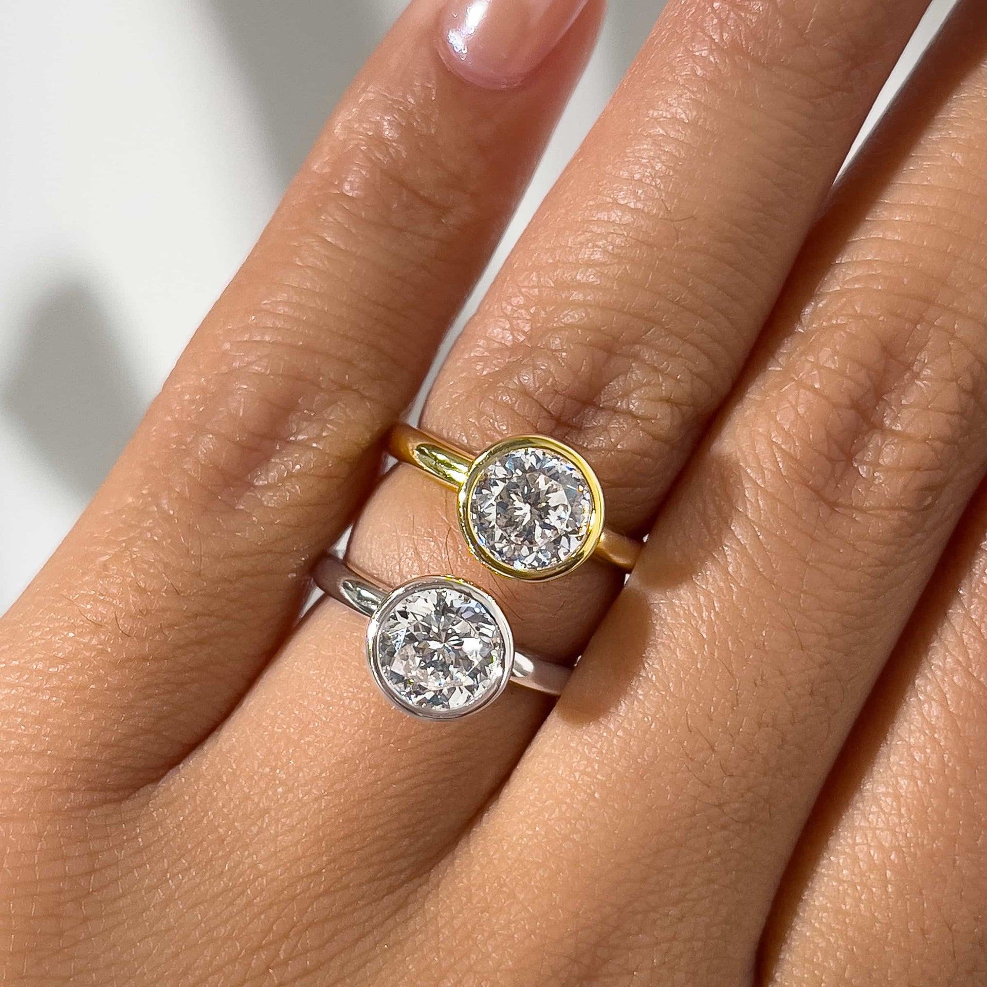 two bezel egagement rings in silver and gold with round cut center stones on model's finger 
