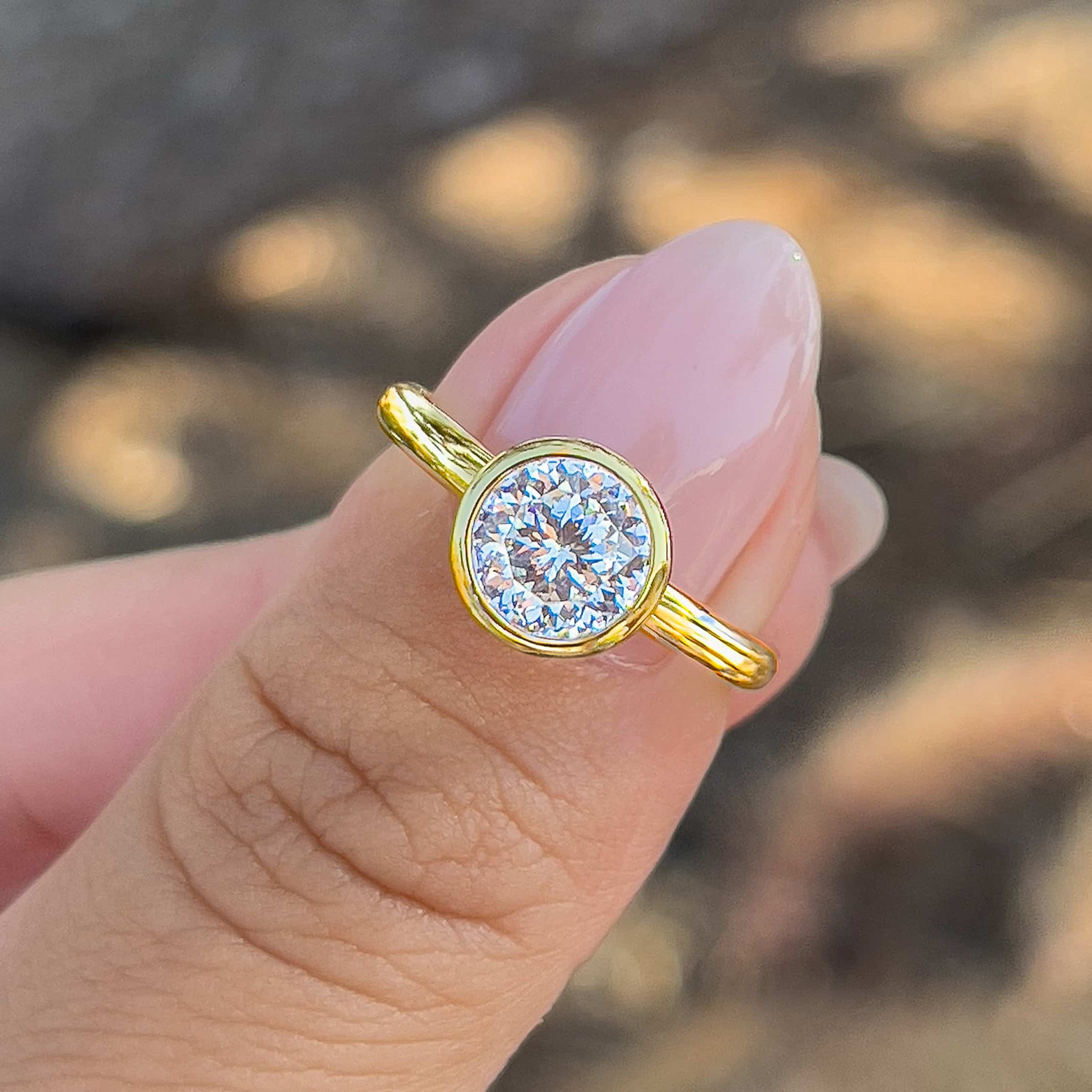 woman with pink nails holding a gold 1.25 carat round cut bezel engagement ring on her thumb