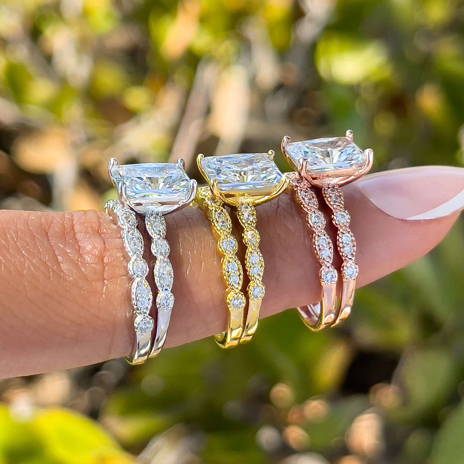 a vintage 1.5 carat radiant cut wedding ring set shown in silver, gold, and rose gold on model with french tip nails in front of greenery