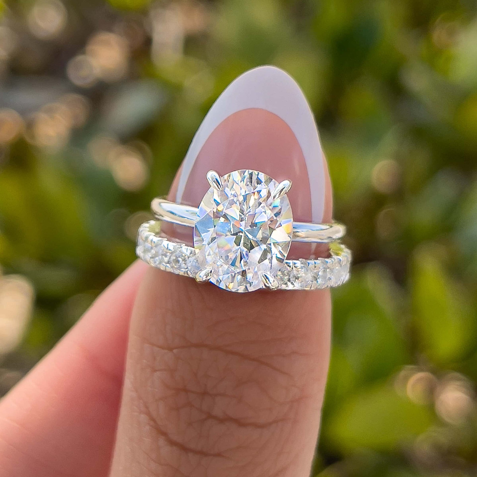 silver 3 carat oval cut solitaire and pavé 
eternity wedding band paired together to create a stunning, simple wedding set on model with french tip nails in front of greenery