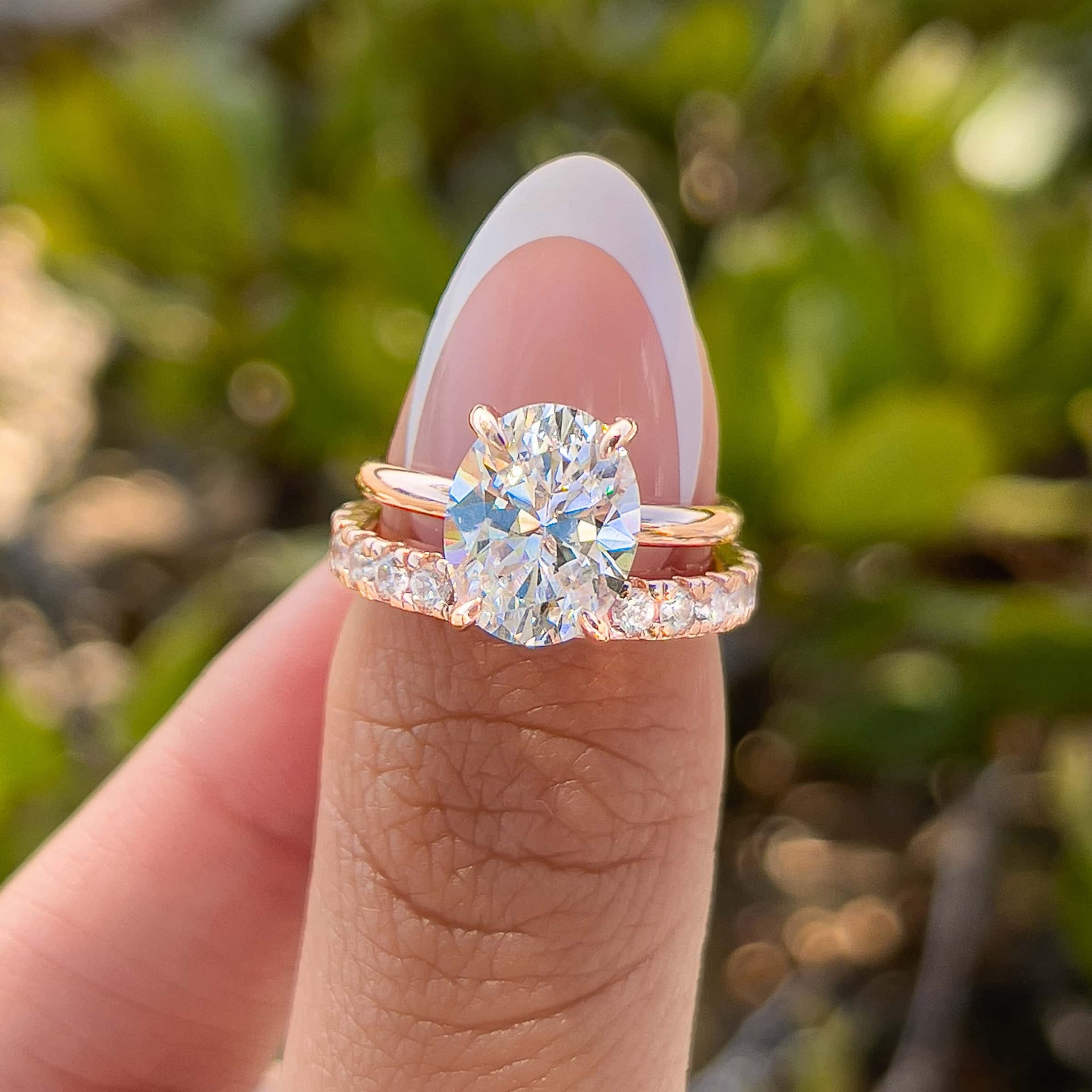 rose gold 3 carat oval cut solitaire and pavé eternity wedding band paired together to create a stunning, simple wedding set on model with french tip nails in front of greenery